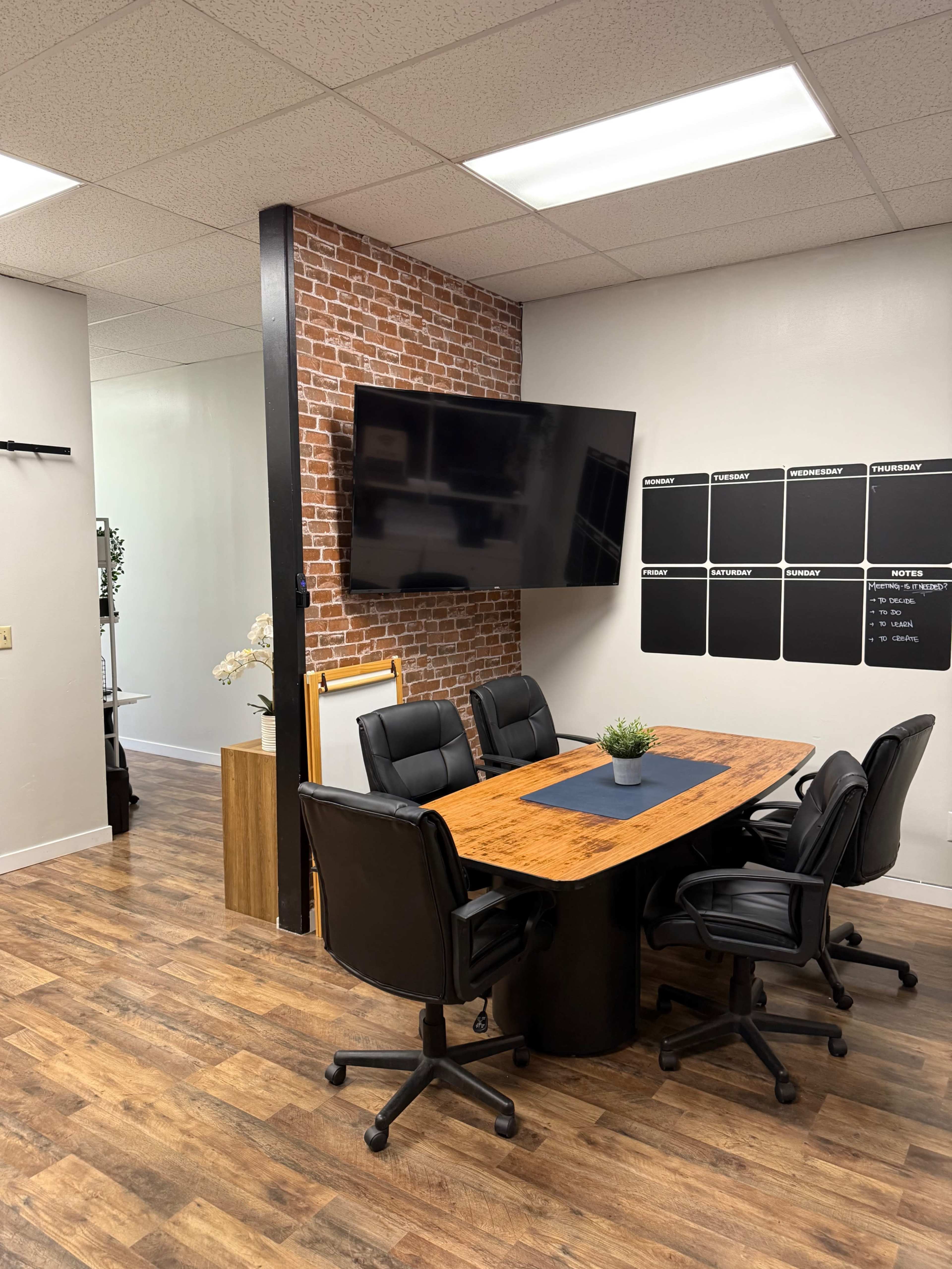 The image shows a conference room with a wooden table and four black chairs, a large wall-mounted television, and a brick accent wall.