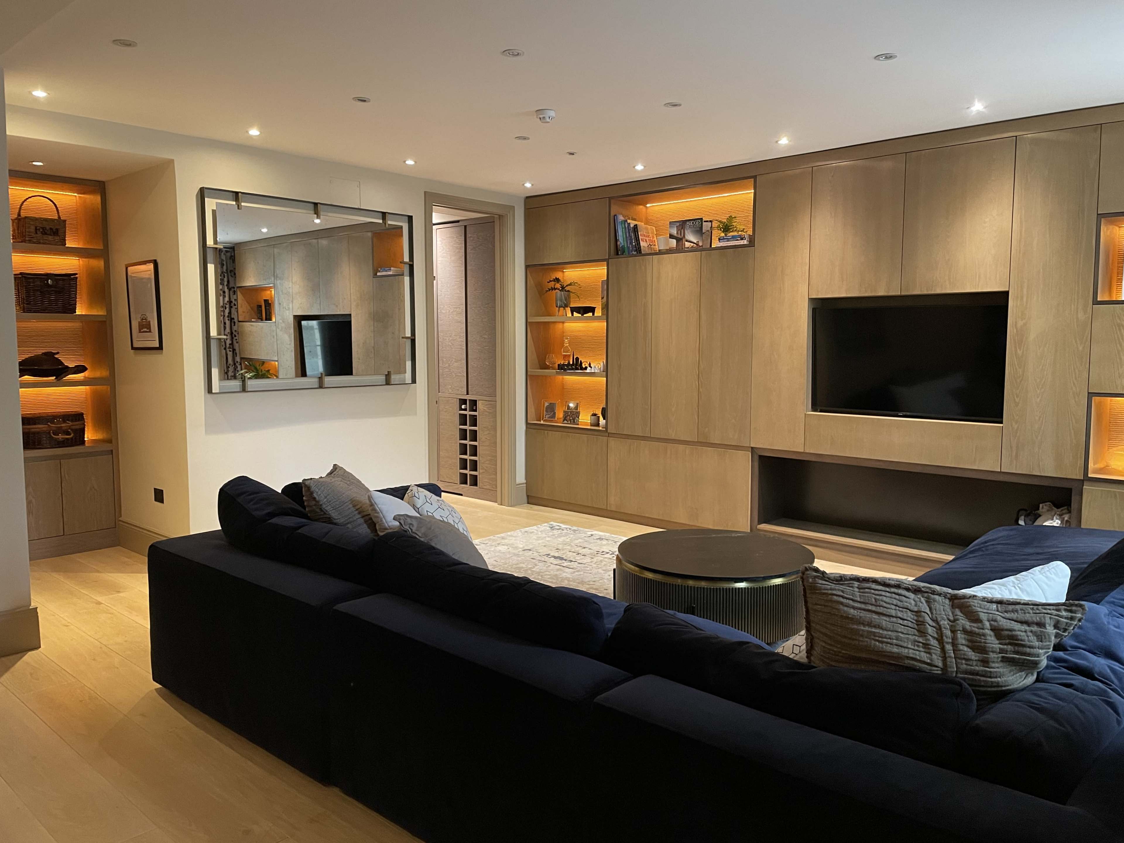 The image shows a modern living room featuring a large dark blue sofa, a coffee table, and wooden shelving with backlighting, along with a television mounted on the wall.