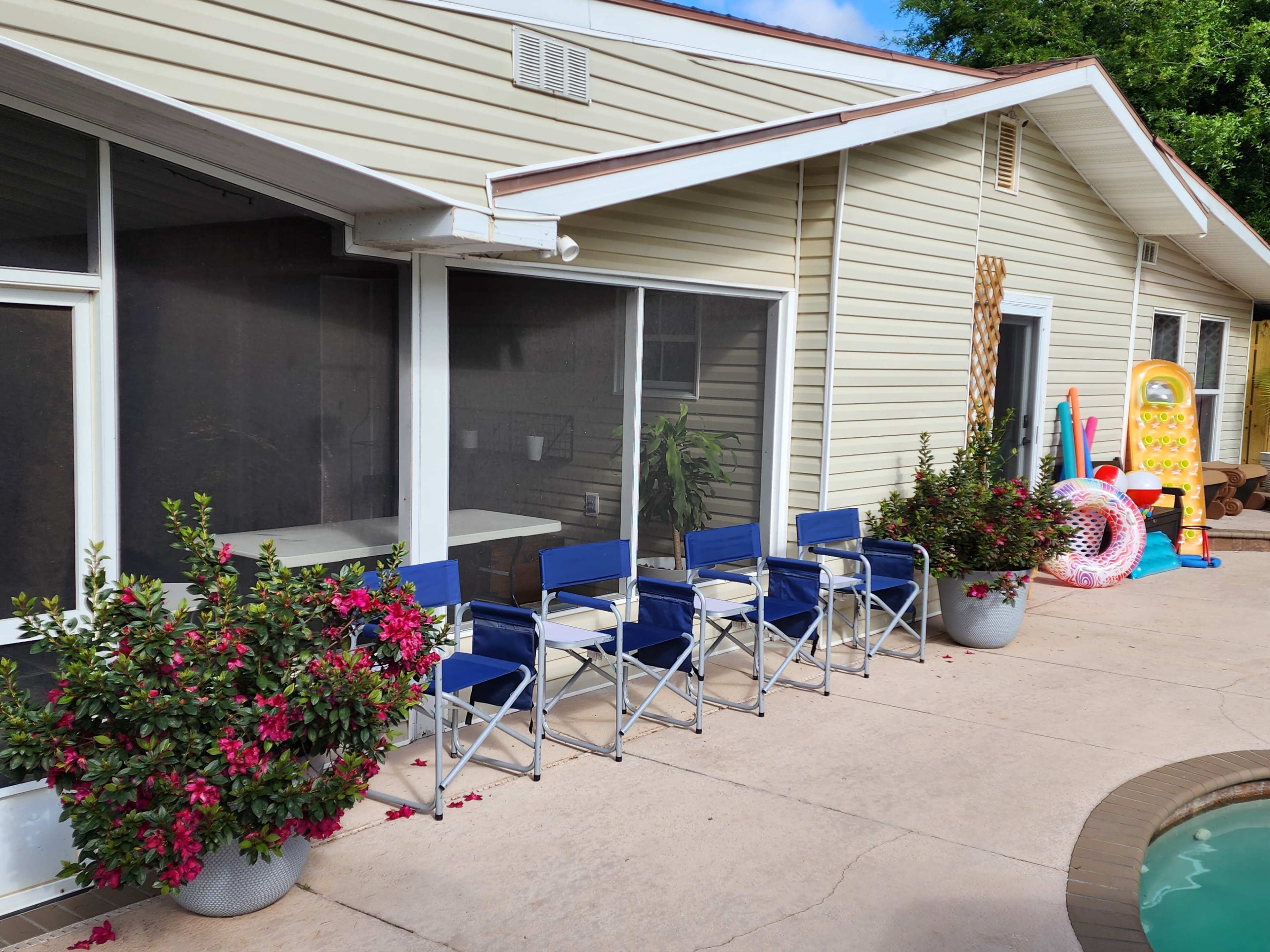 The image shows a patio area beside a swimming pool, featuring a row of blue and white folding chairs and potted plants with pink flowers.