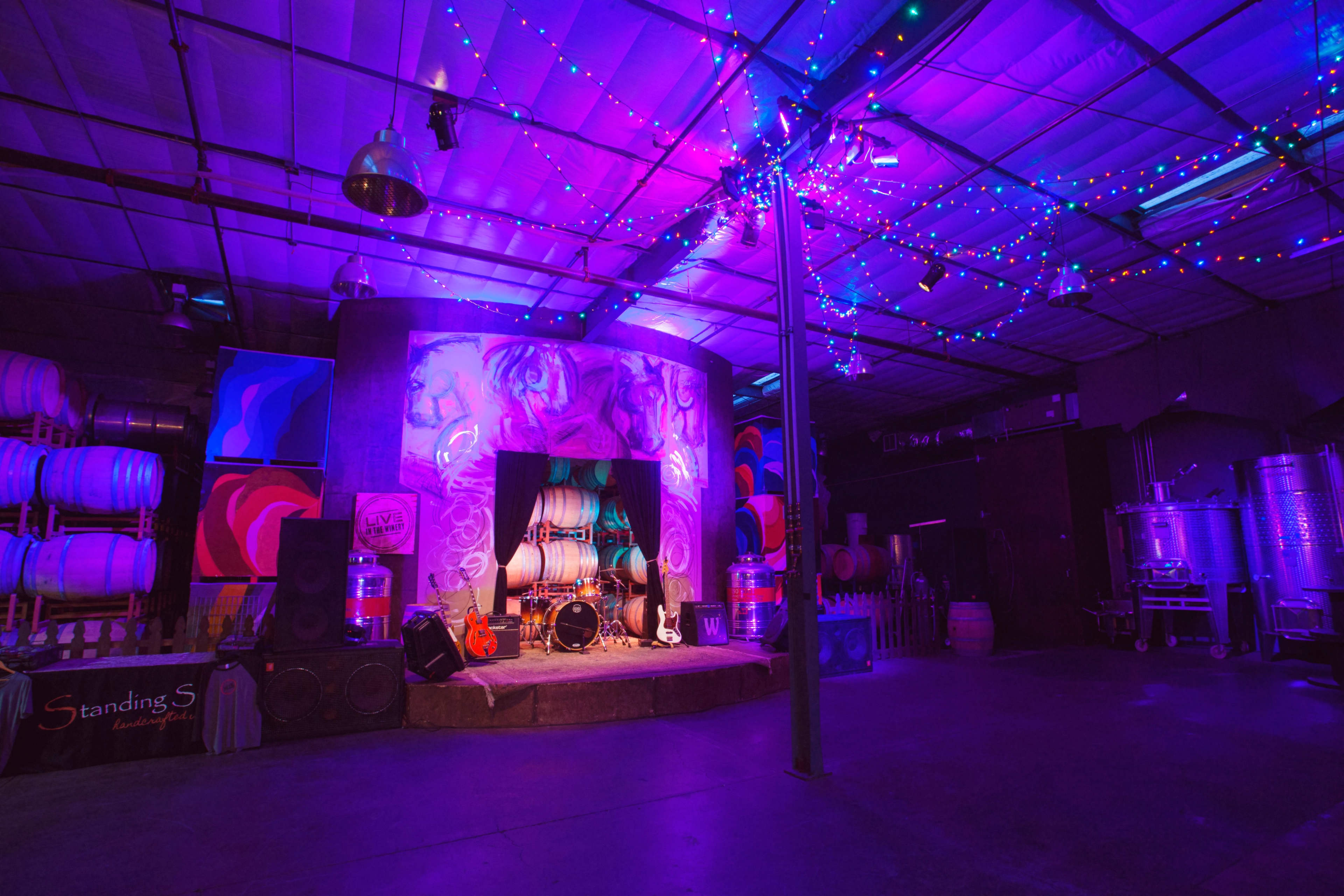 A dimly lit event space with colorful lights, featuring a decorated stage, musical instruments, and wine barrels in the background.