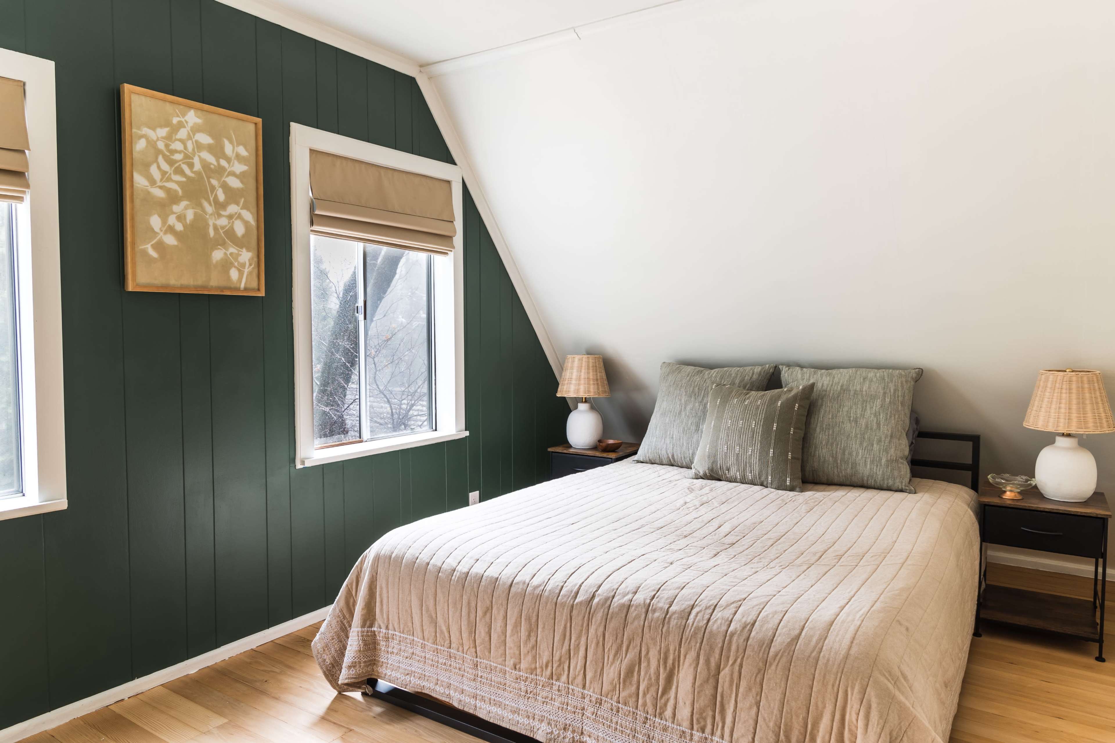 The image shows a minimalist bedroom featuring a bed with neutral-colored bedding against a dark green wall and two windows with shades.