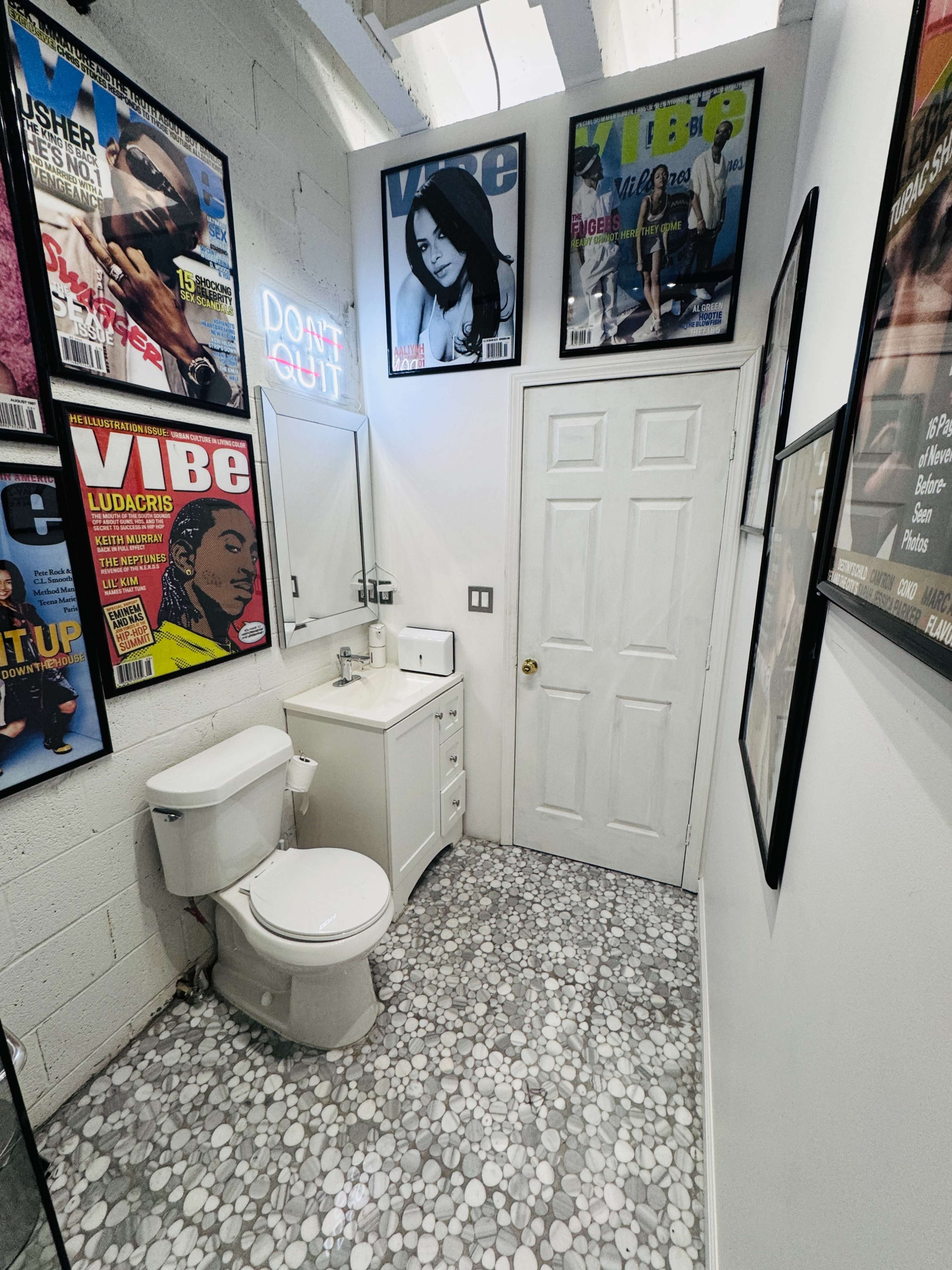 The image shows a small bathroom featuring a toilet, sink with a mirror, and walls adorned with framed magazine covers, all set against a pebble-patterned floor.