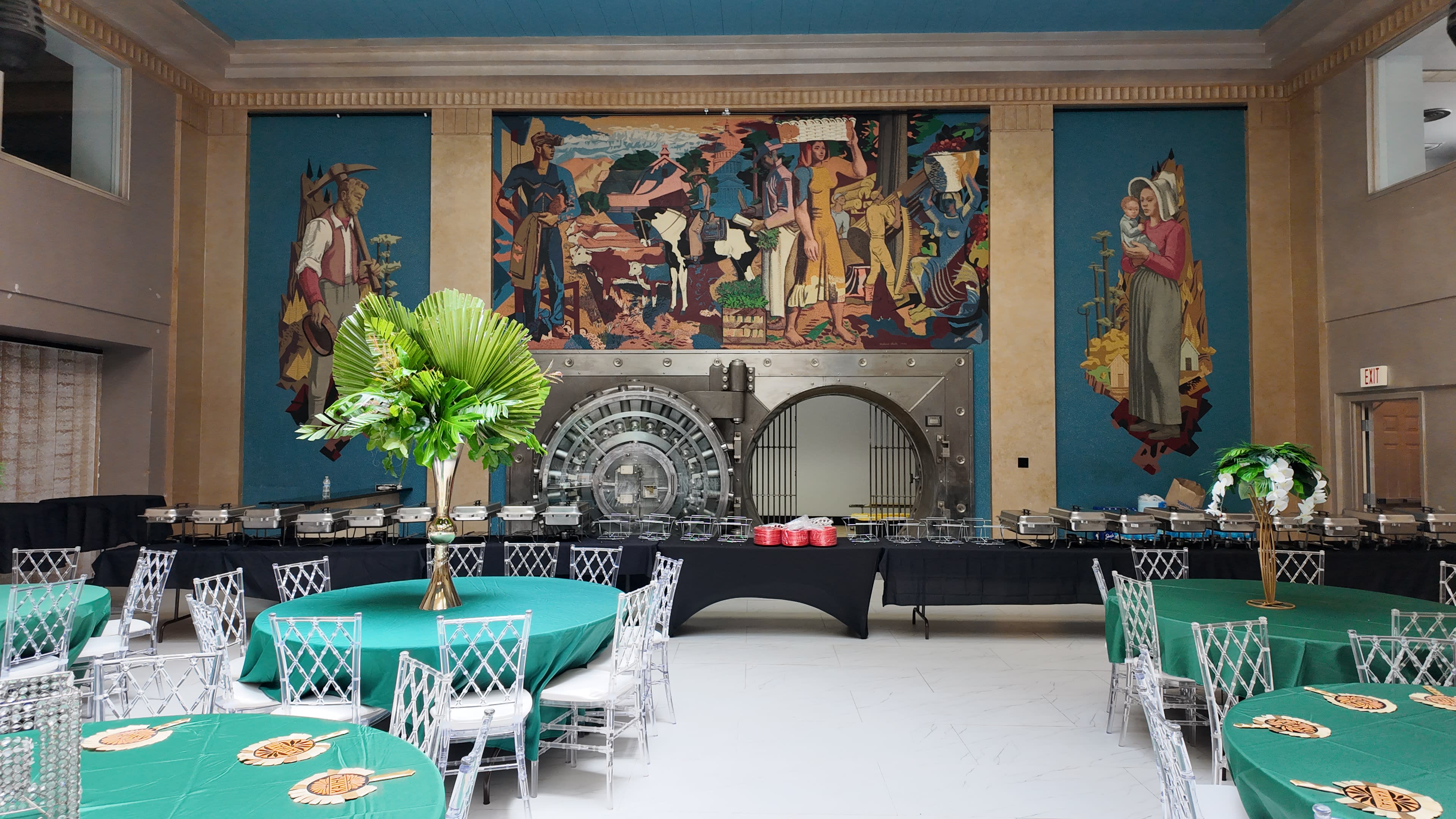 A large banquet hall features round tables with green tablecloths and a buffet setup against a backdrop of colorful murals and a prominent vault door.
