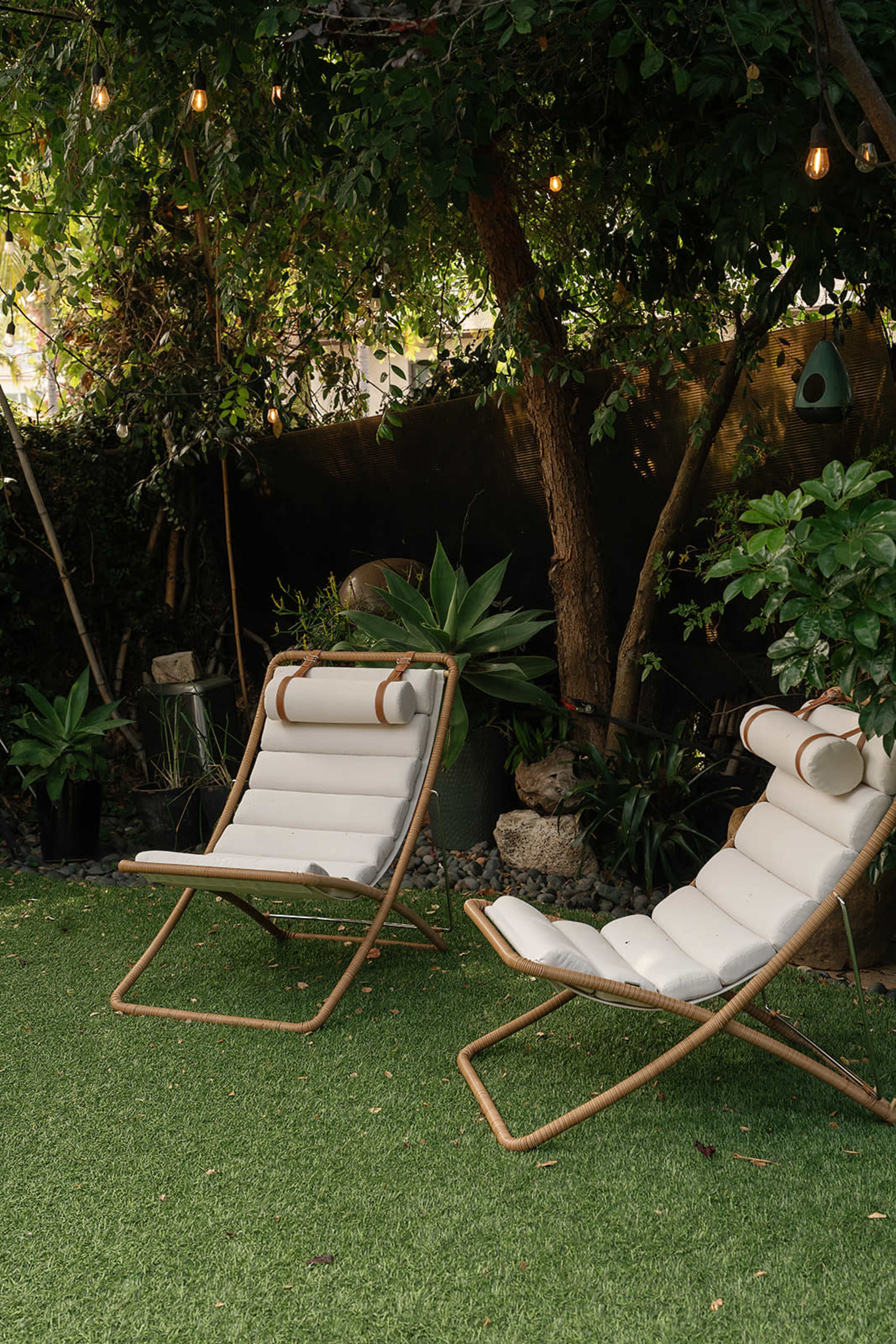 Two lounge chairs are positioned on a grassy area beneath trees adorned with string lights, surrounded by various plants.