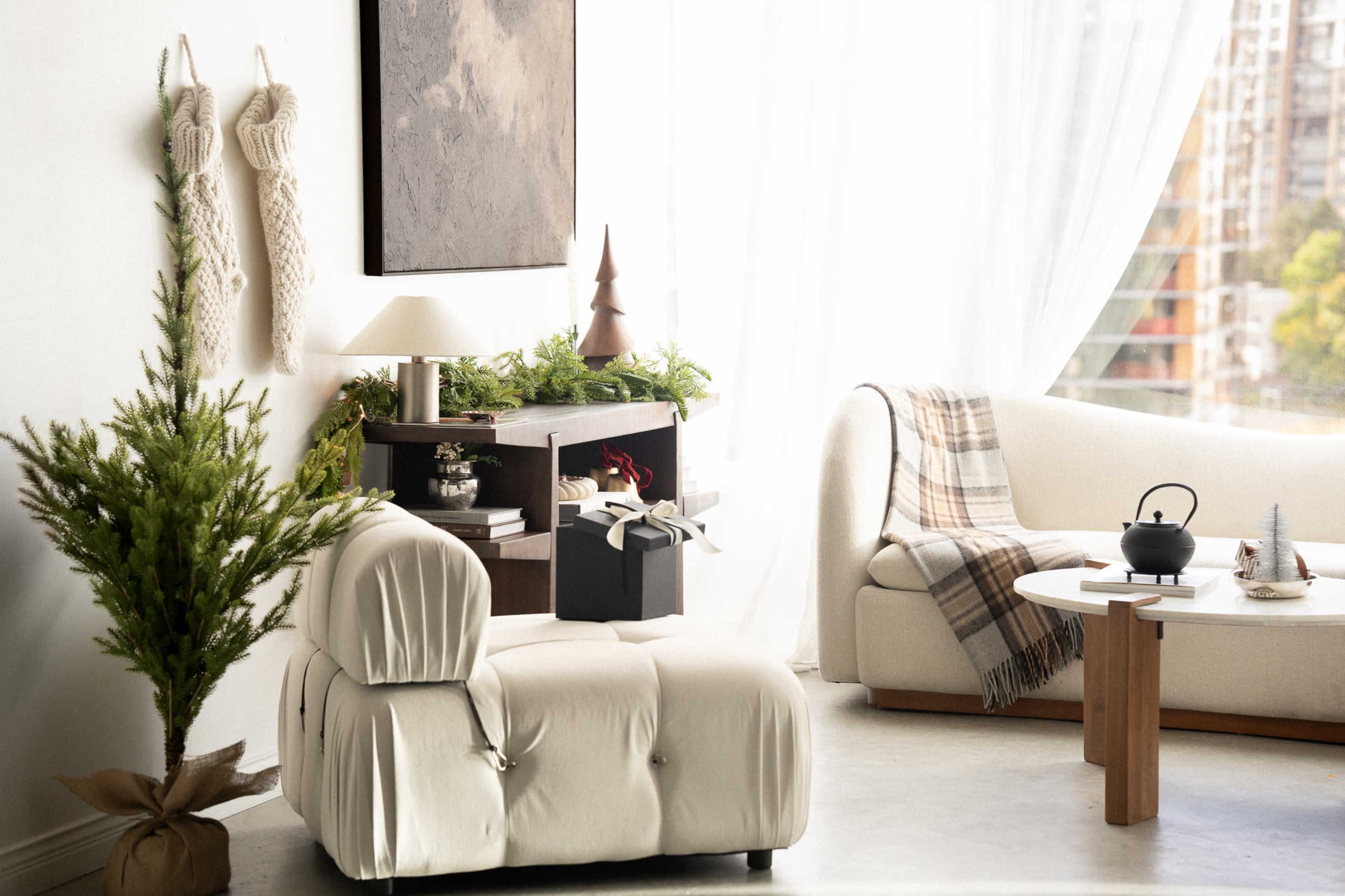 The image depicts a cozy living room scene featuring a light-colored chair, decorative elements like a small Christmas tree, and a table with a black teapot and gift box.