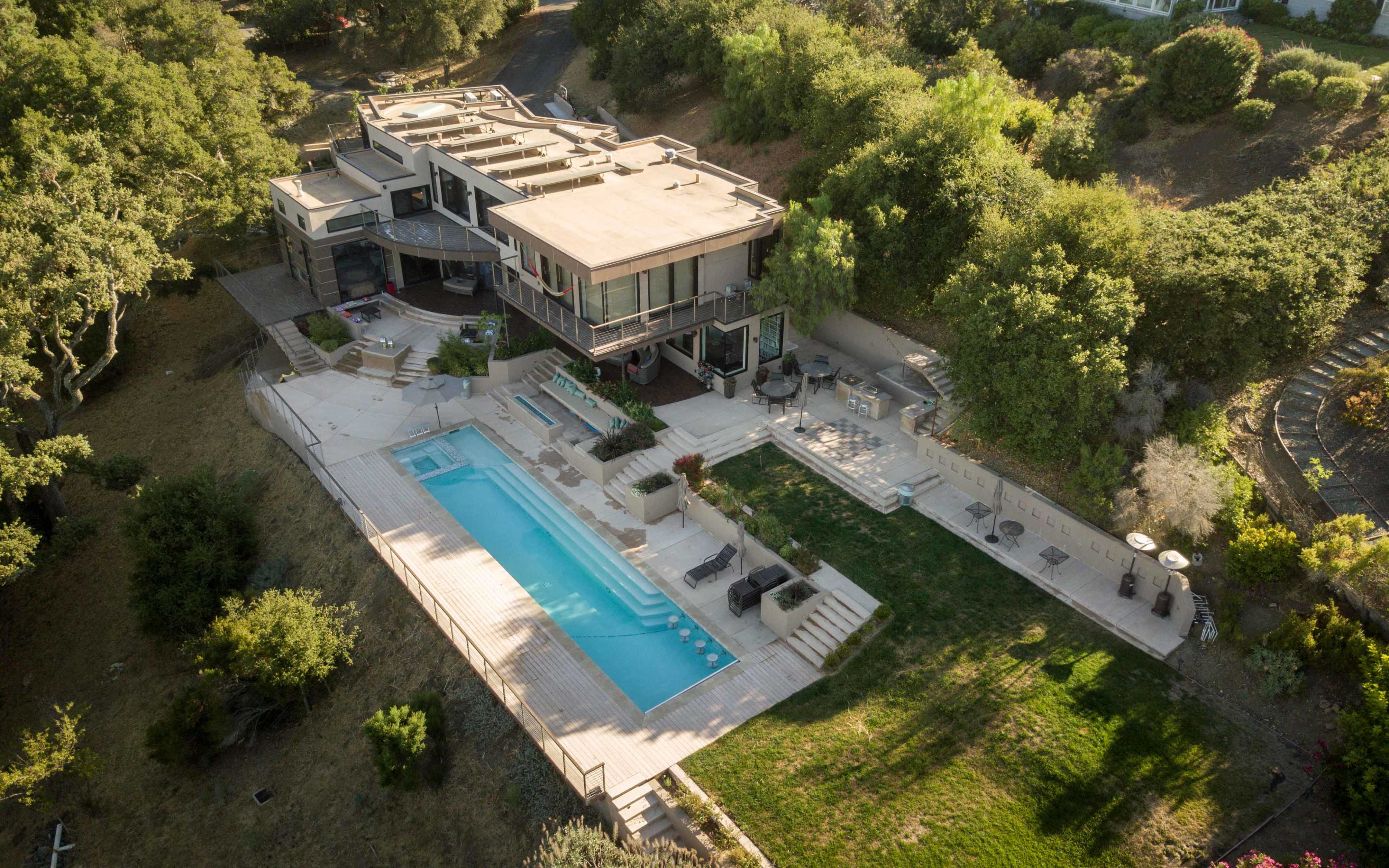 The image shows a modern house with a flat roof, featuring a large swimming pool and landscaped outdoor spaces.