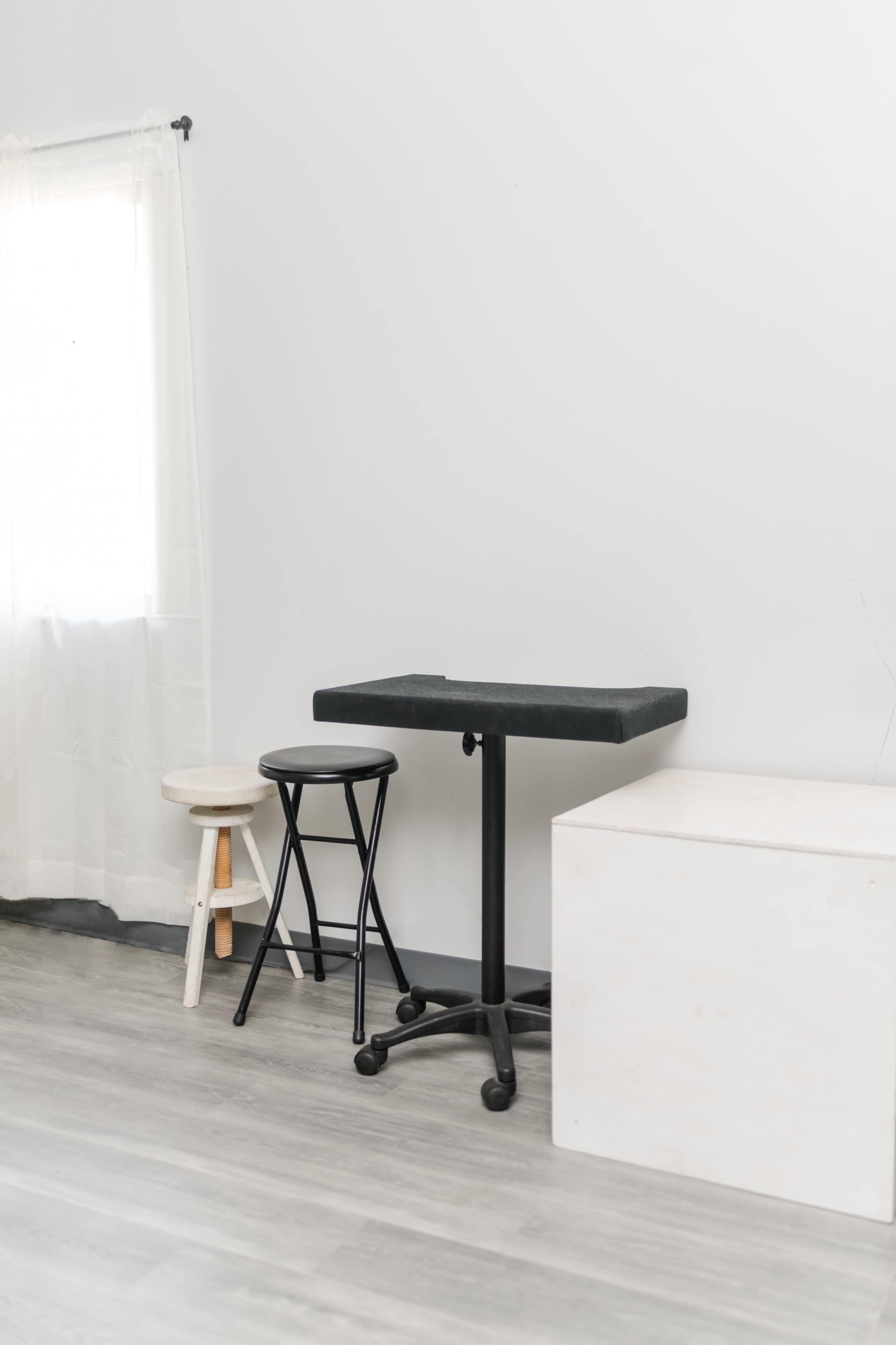A minimalist interior with a black table on wheels, two stools of different heights, and a white storage box against a plain wall.