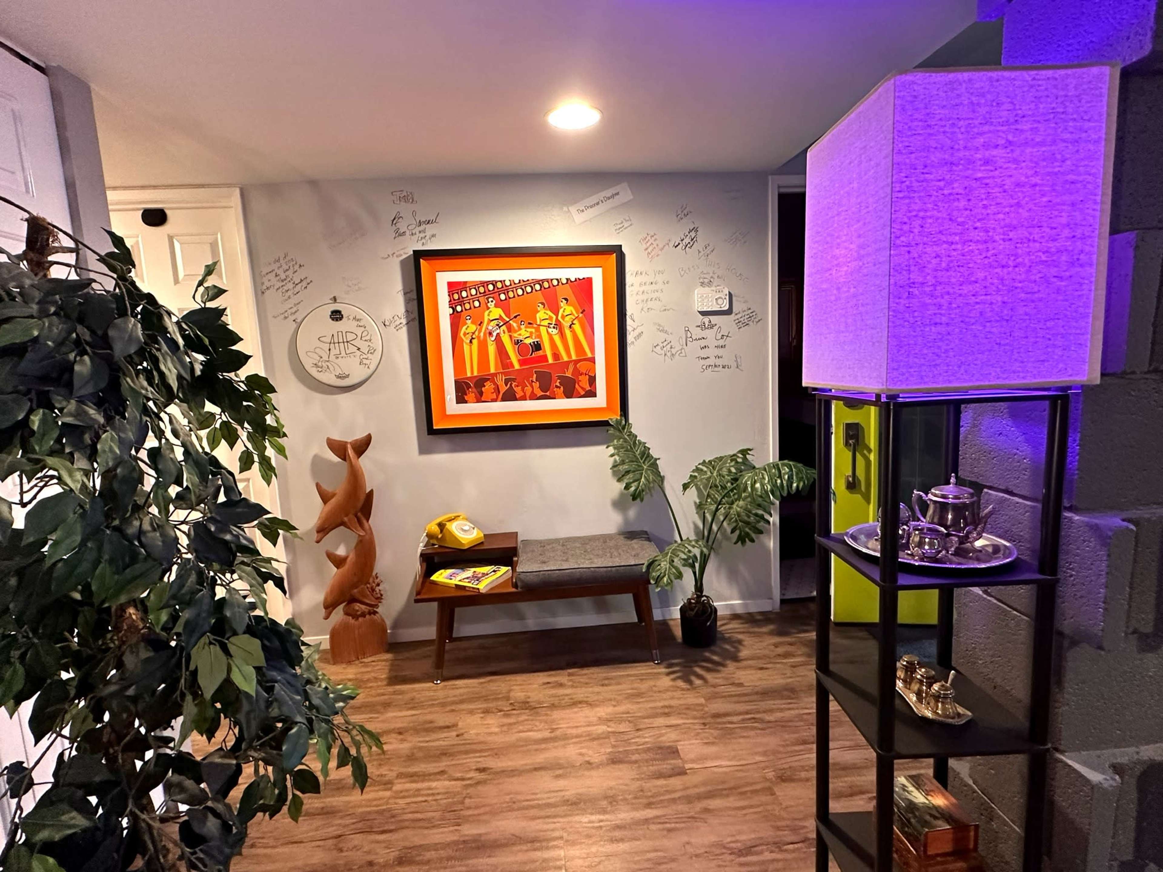 A cozy room featuring a colorful framed artwork, a bench, a potted plant, and a lit lamp beside a decorative shelf.