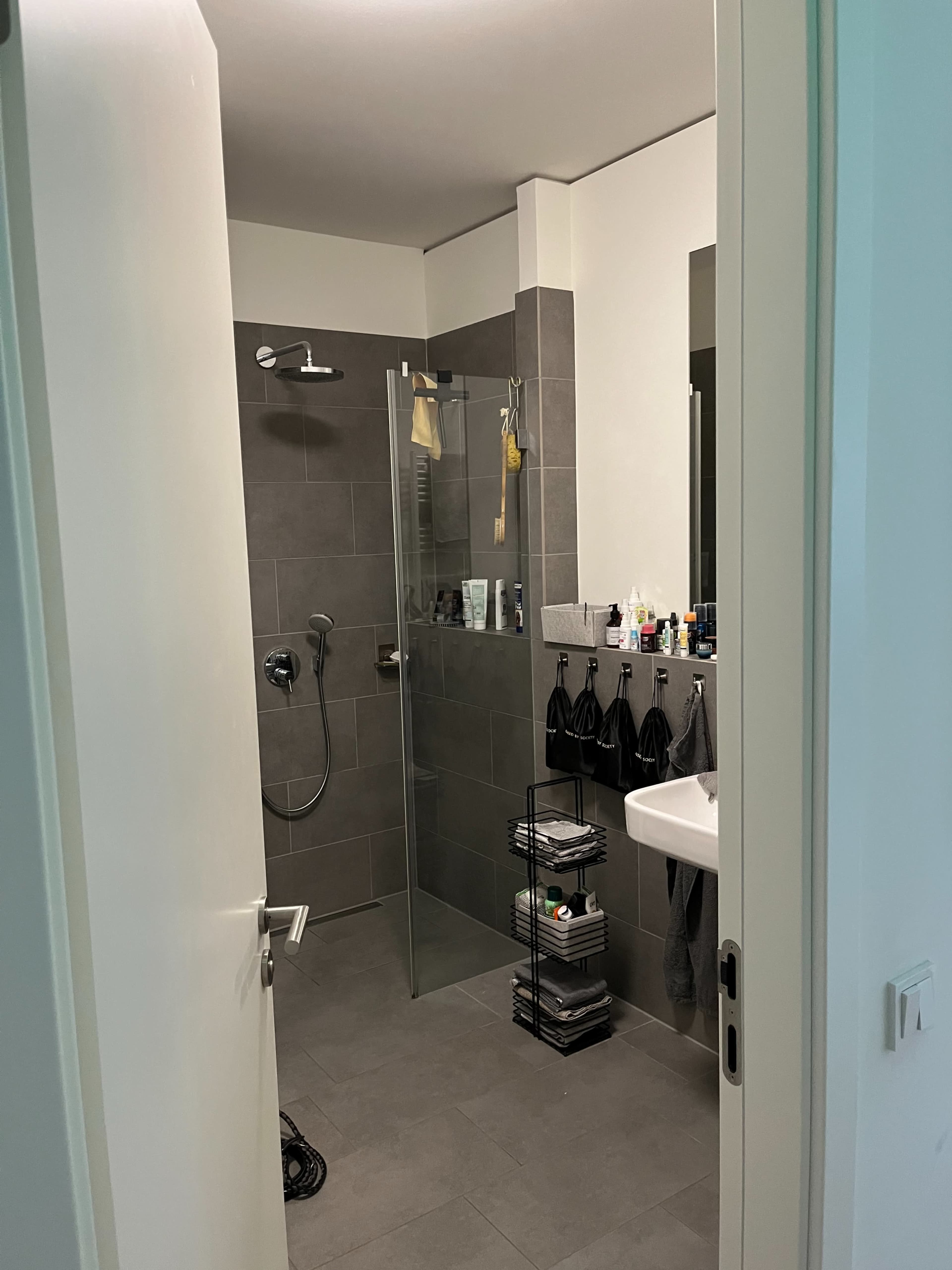 A modern bathroom features a glass shower, a sink with toiletries, and shelves stacked with towels and toiletries.