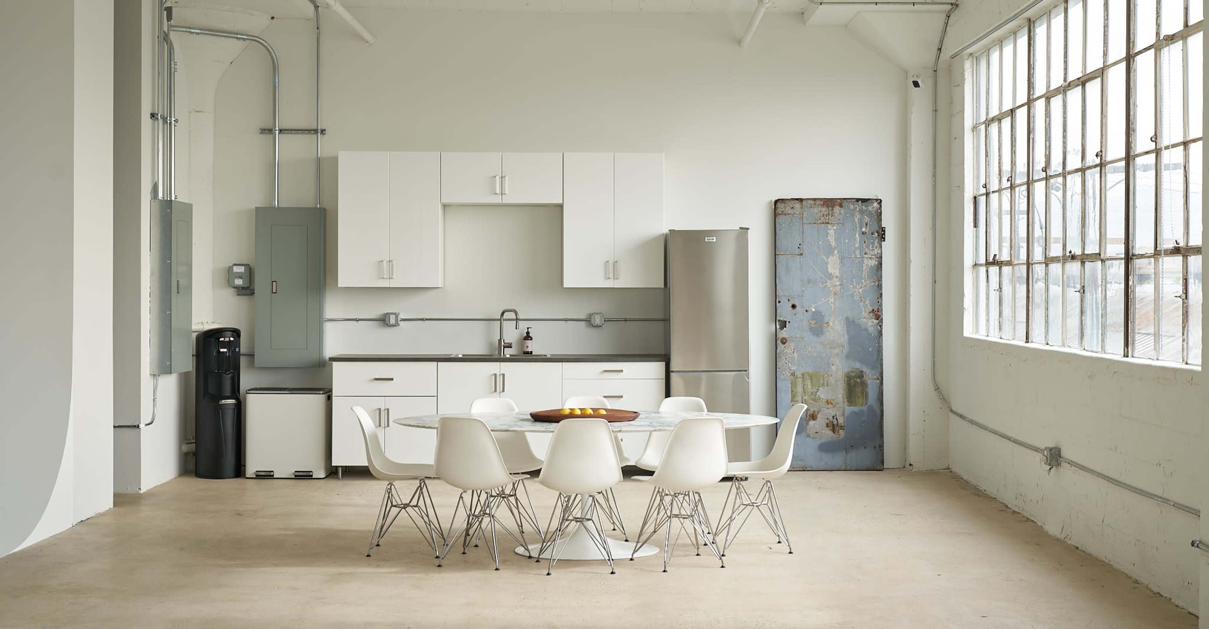 The image shows a spacious kitchen area featuring a round dining table surrounded by white chairs, with a sink, refrigerator, and cabinets along one wall.