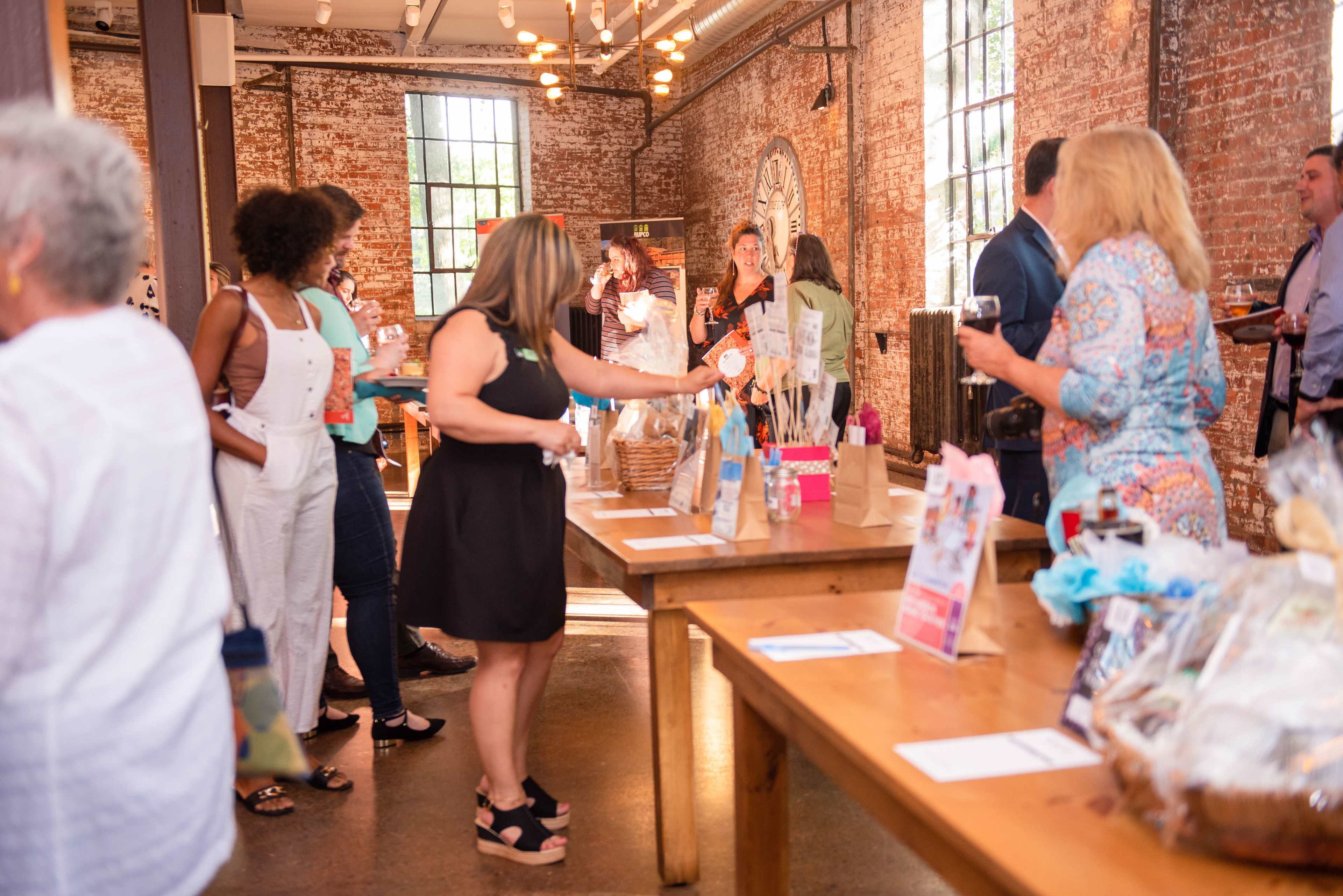 A group of people socializes and mingles at a gathering in a warmly lit, rustic venue with exposed brick walls and wooden tables.