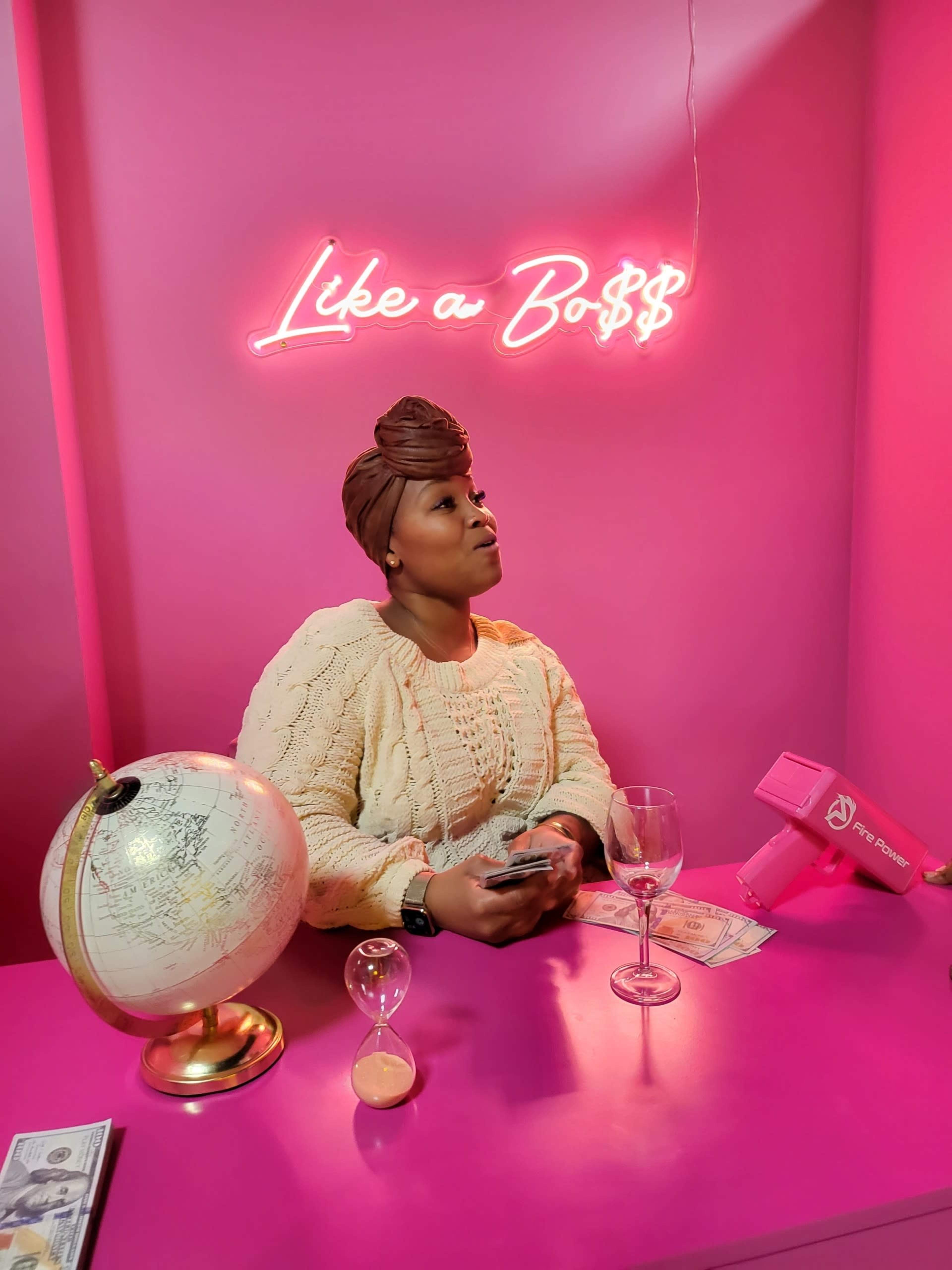 A woman sits at a pink table holding cash, with a globe and an hourglass nearby, while a neon sign above her reads "Like a Bo$$."
