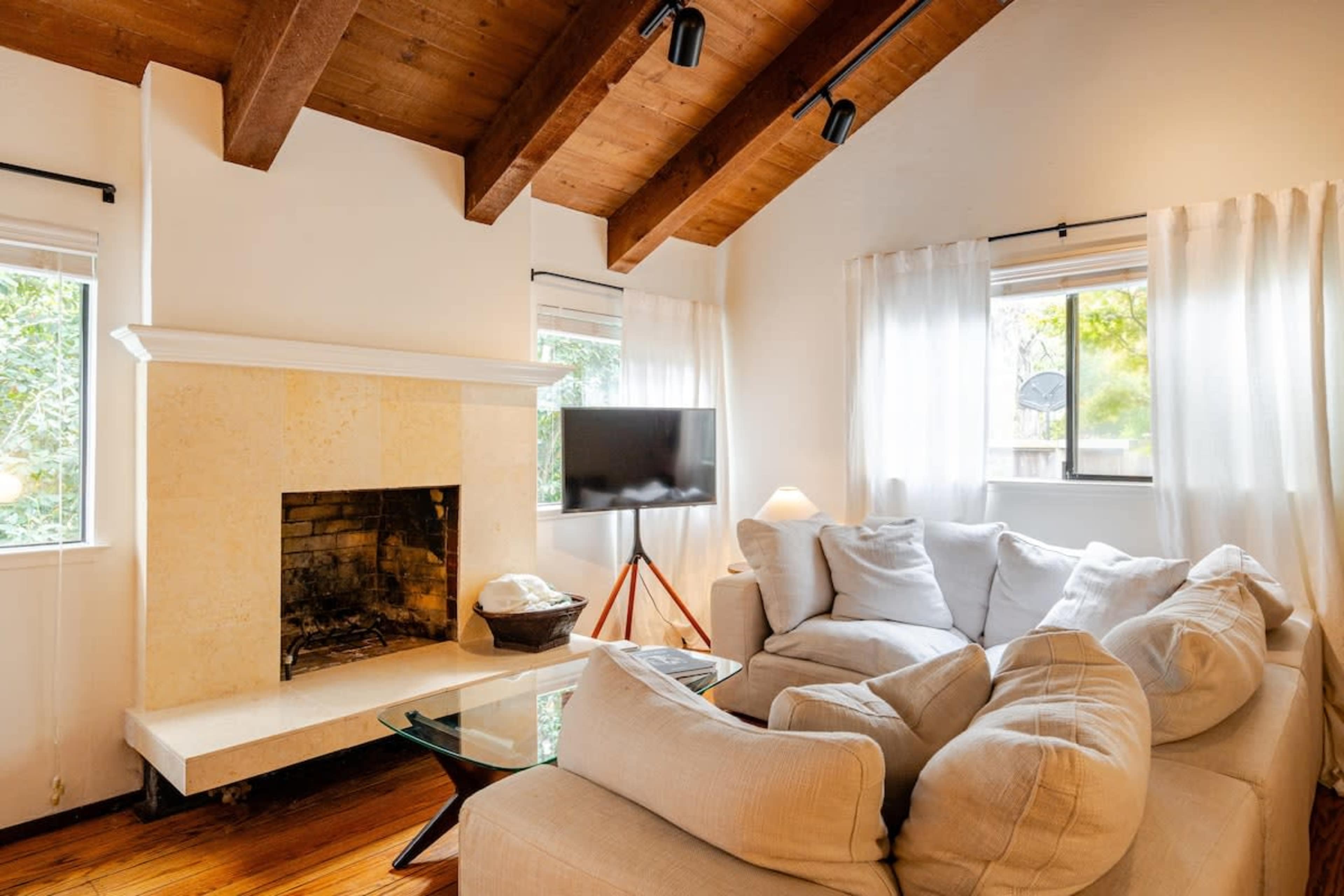 The image shows a cozy living room featuring a white sectional sofa, a glass coffee table, and a stone fireplace, with large windows allowing natural light to enter.
