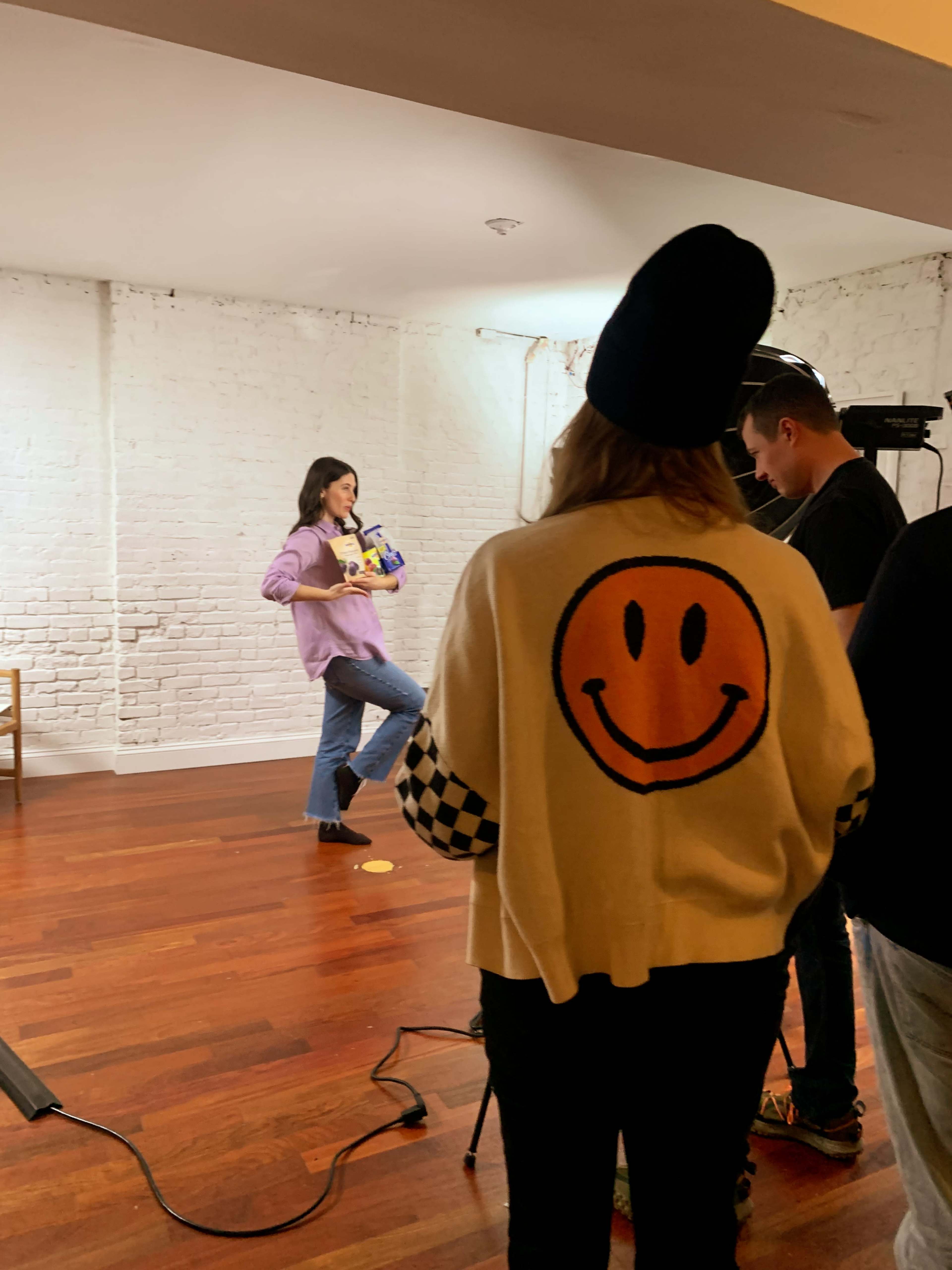 A person poses with a box on one leg while another individual in a beanie and a smiling sweater watches, and a camera is set up nearby in a studio setting.