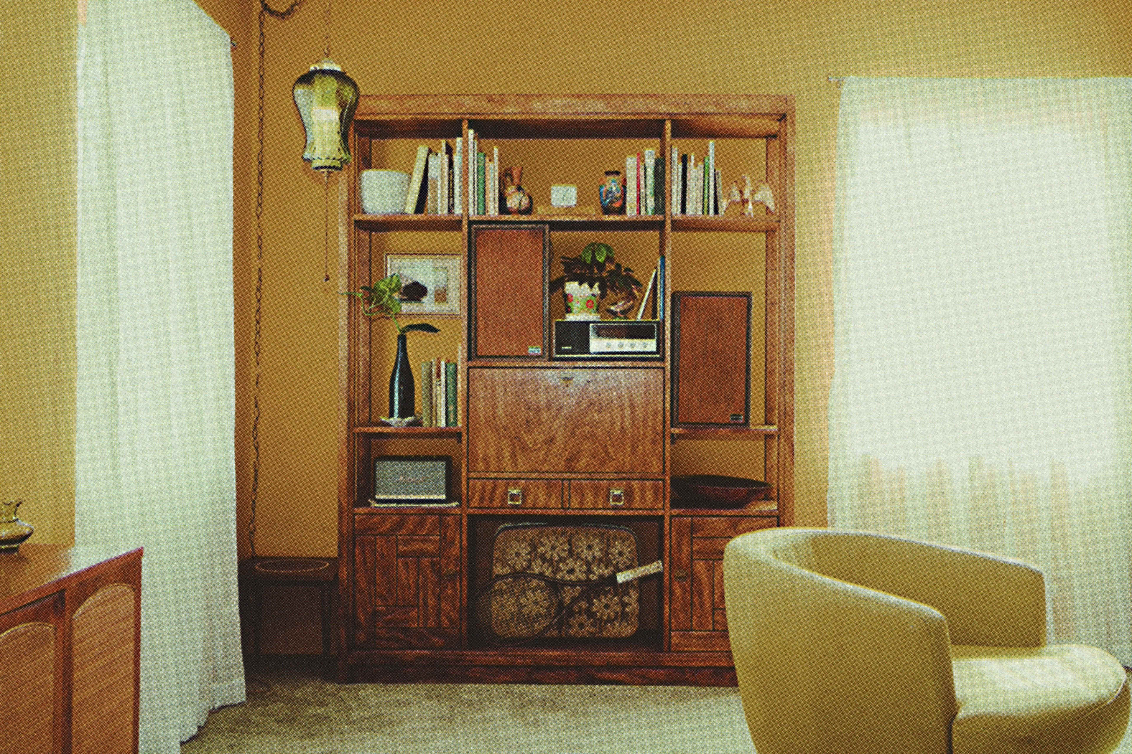 A wooden shelving unit filled with books and decorative items stands against a wall in a room with beige walls and a light-colored chair.