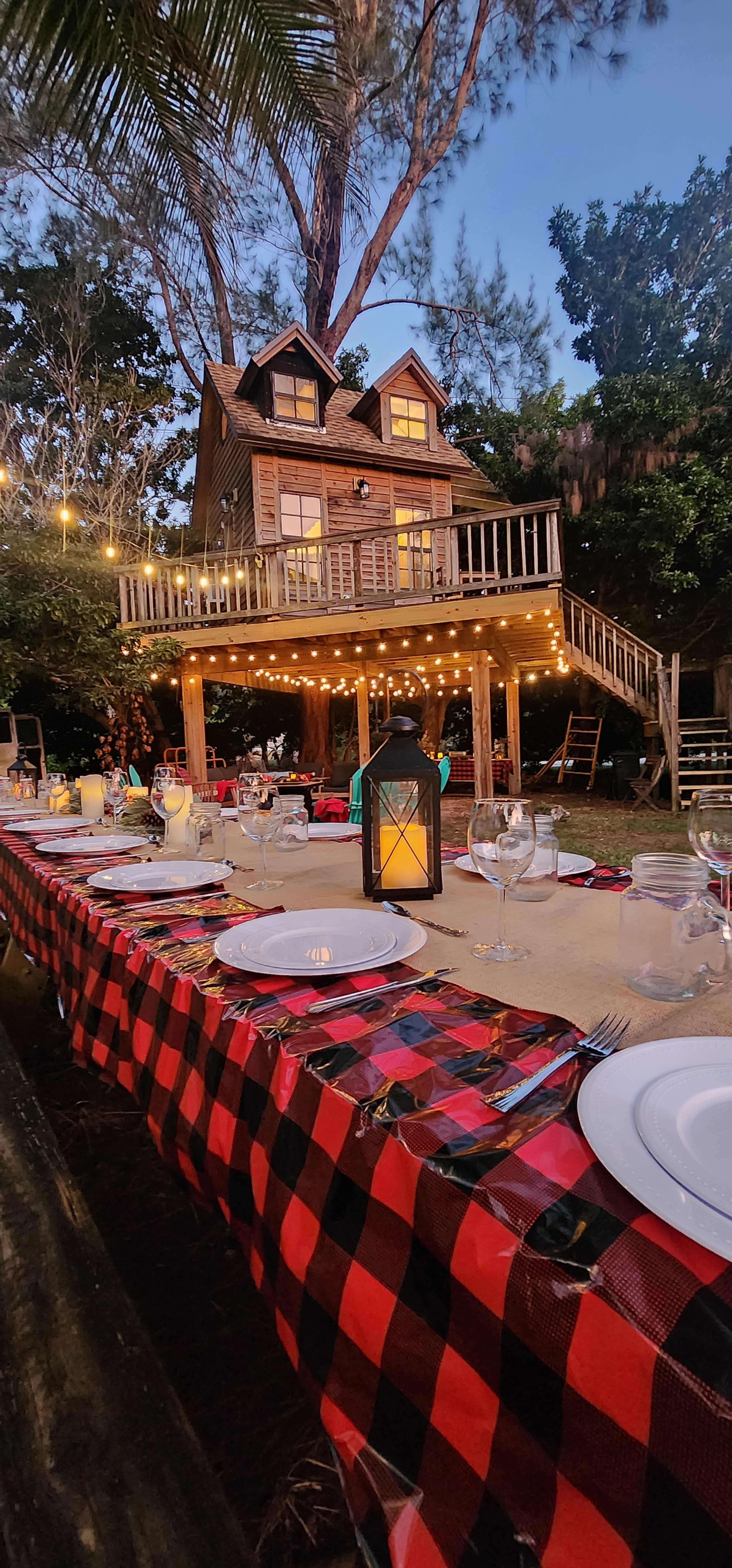 A beautifully set outdoor dining table is positioned in front of a wooden house on stilts, surrounded by trees and illuminated by string lights.