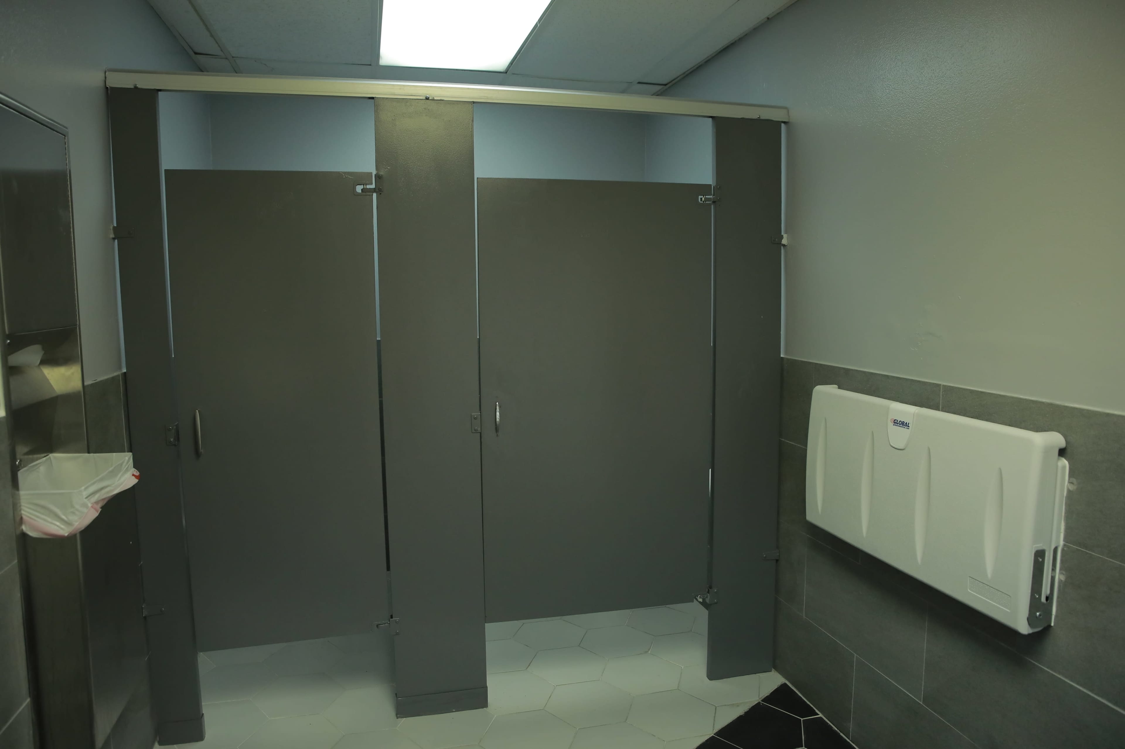 The image shows a bathroom stall area with three gray partitions, a mirror, a trash bin, and a changing table mounted on the wall.