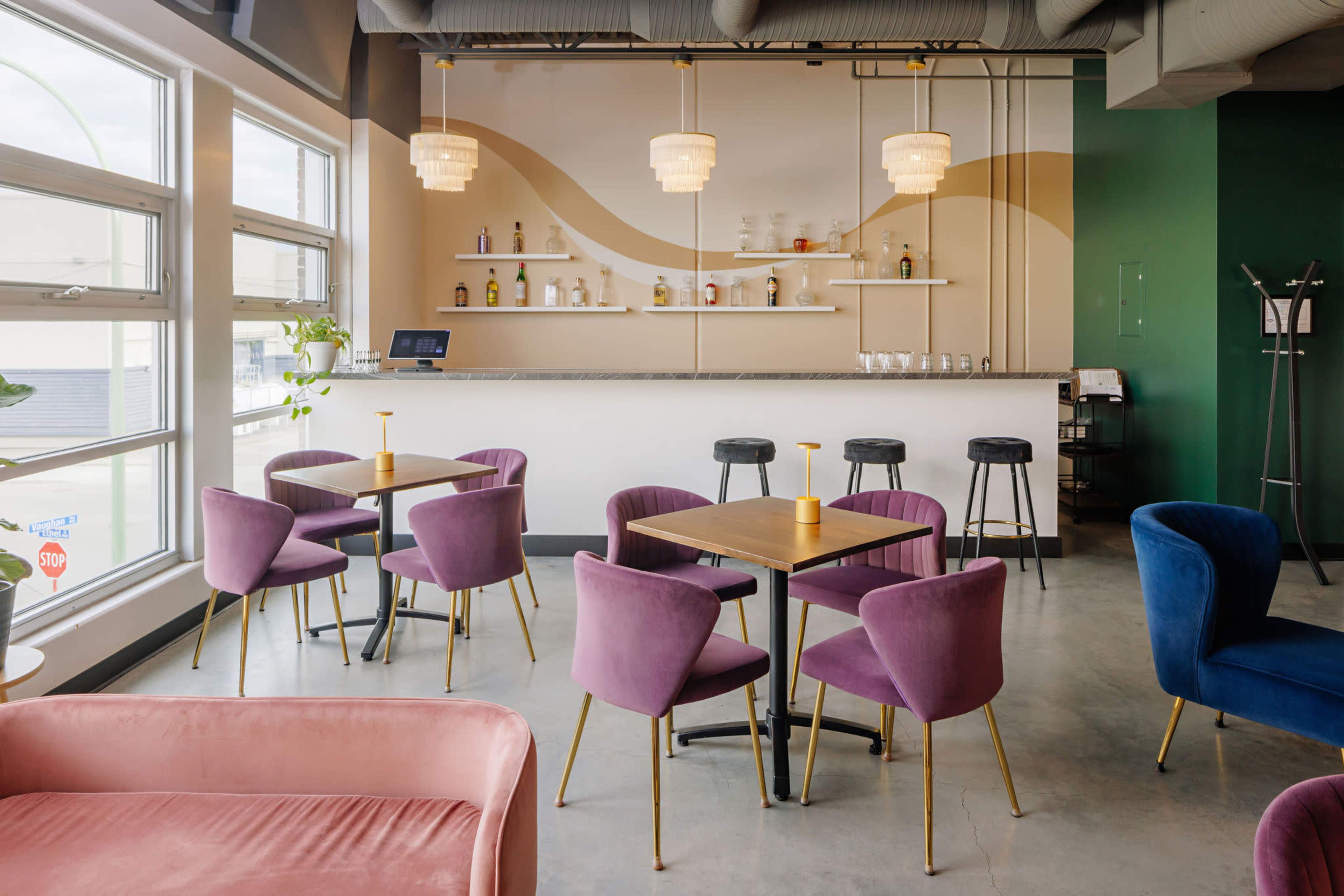 The image shows a modern café interior featuring purple and pink upholstered chairs, wooden tables, and a bar area with shelves of liquor and decorative lighting.