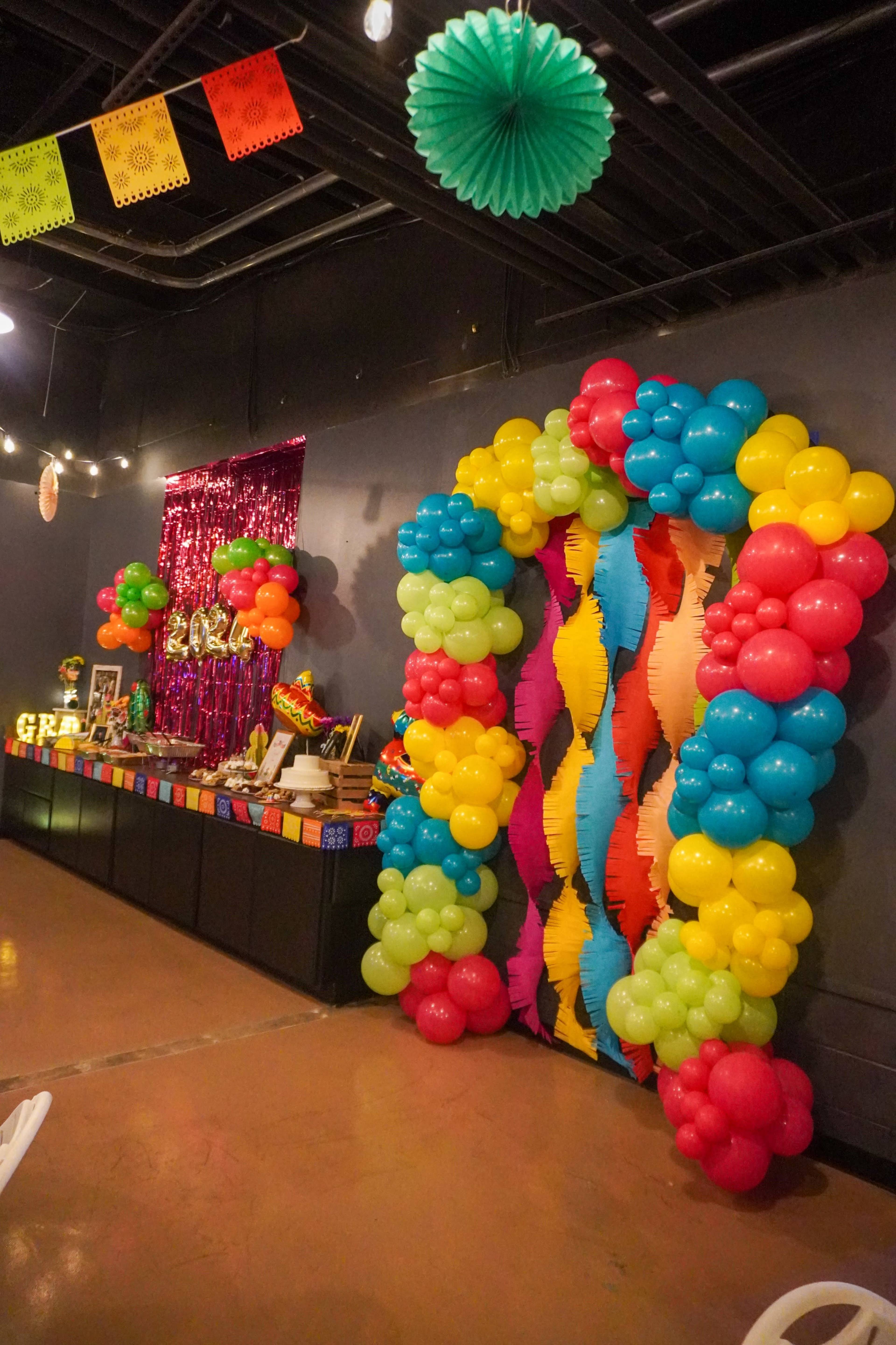 A colorful balloon arch in bright hues frames a decorative display with vibrant table settings and festive banners in a party setting.