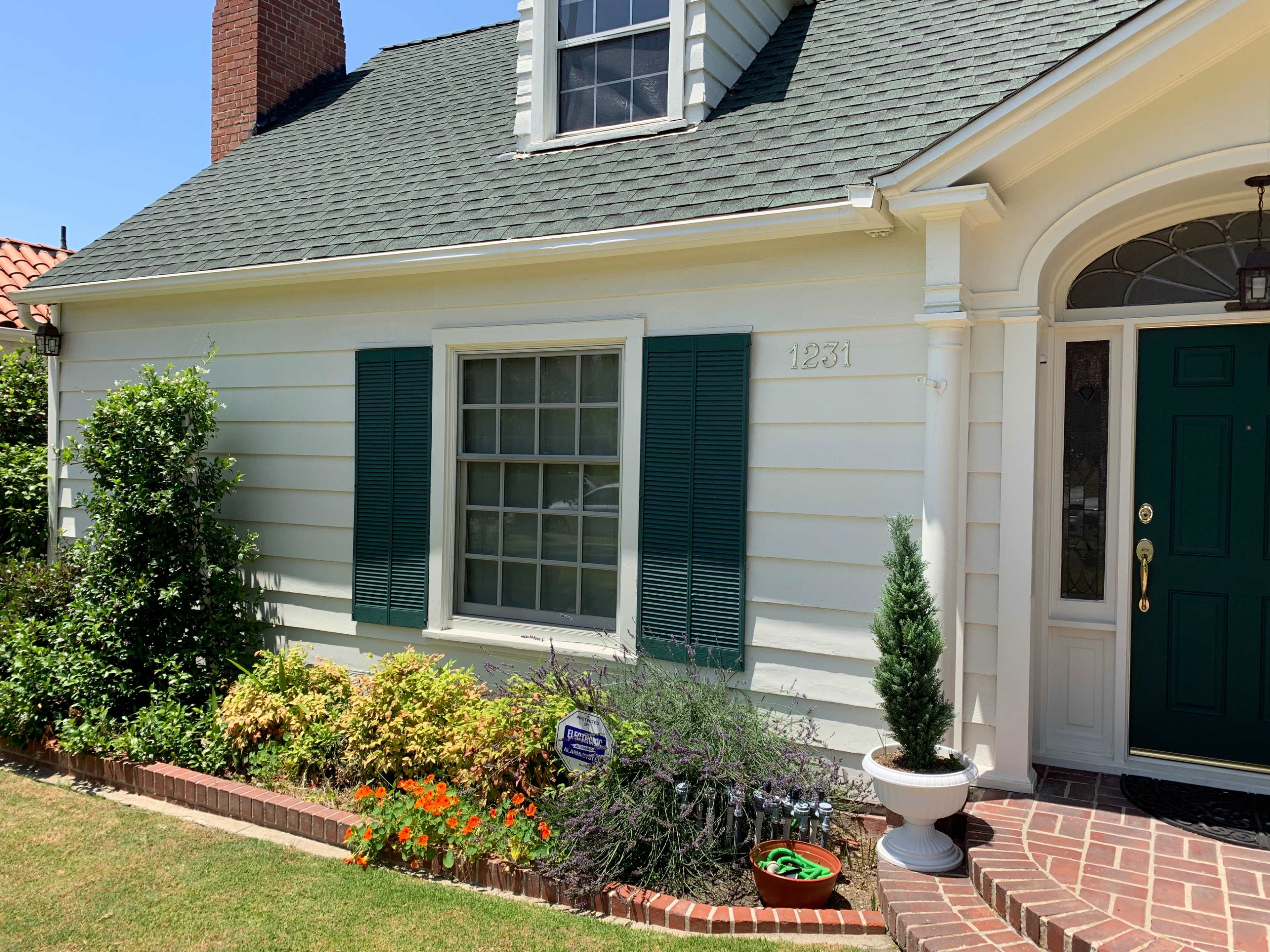 A white house with green shutters, a dark green door, and a neatly landscaped front yard featuring flowers and shrubs.