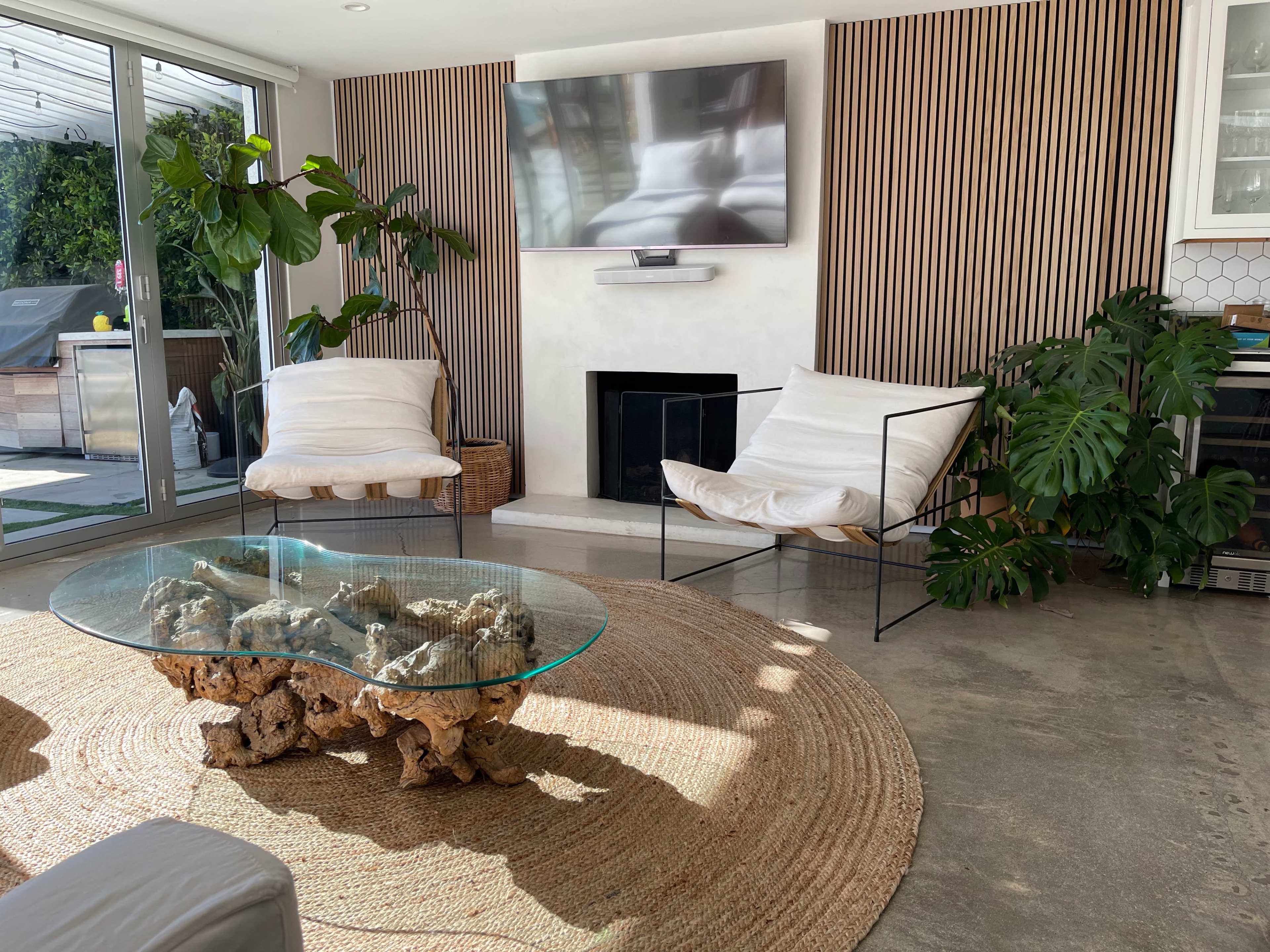 The scene features a modern living room with two chairs, a round rug, a glass coffee table made from natural wood, and a wall-mounted television.