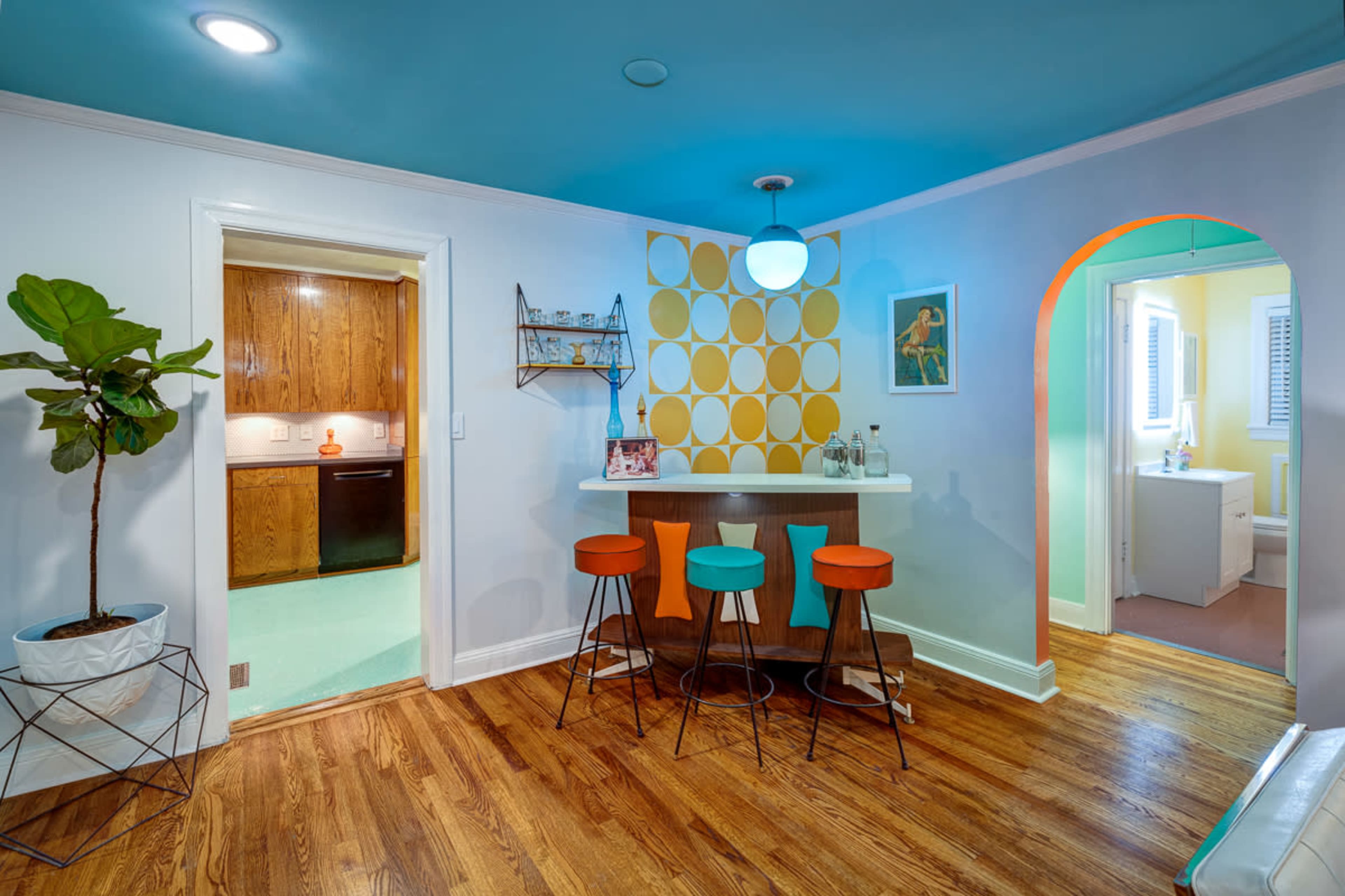 A brightly colored room featuring a small bar area with three stools, a kitchen in the background, and an arched doorway leading to another space.