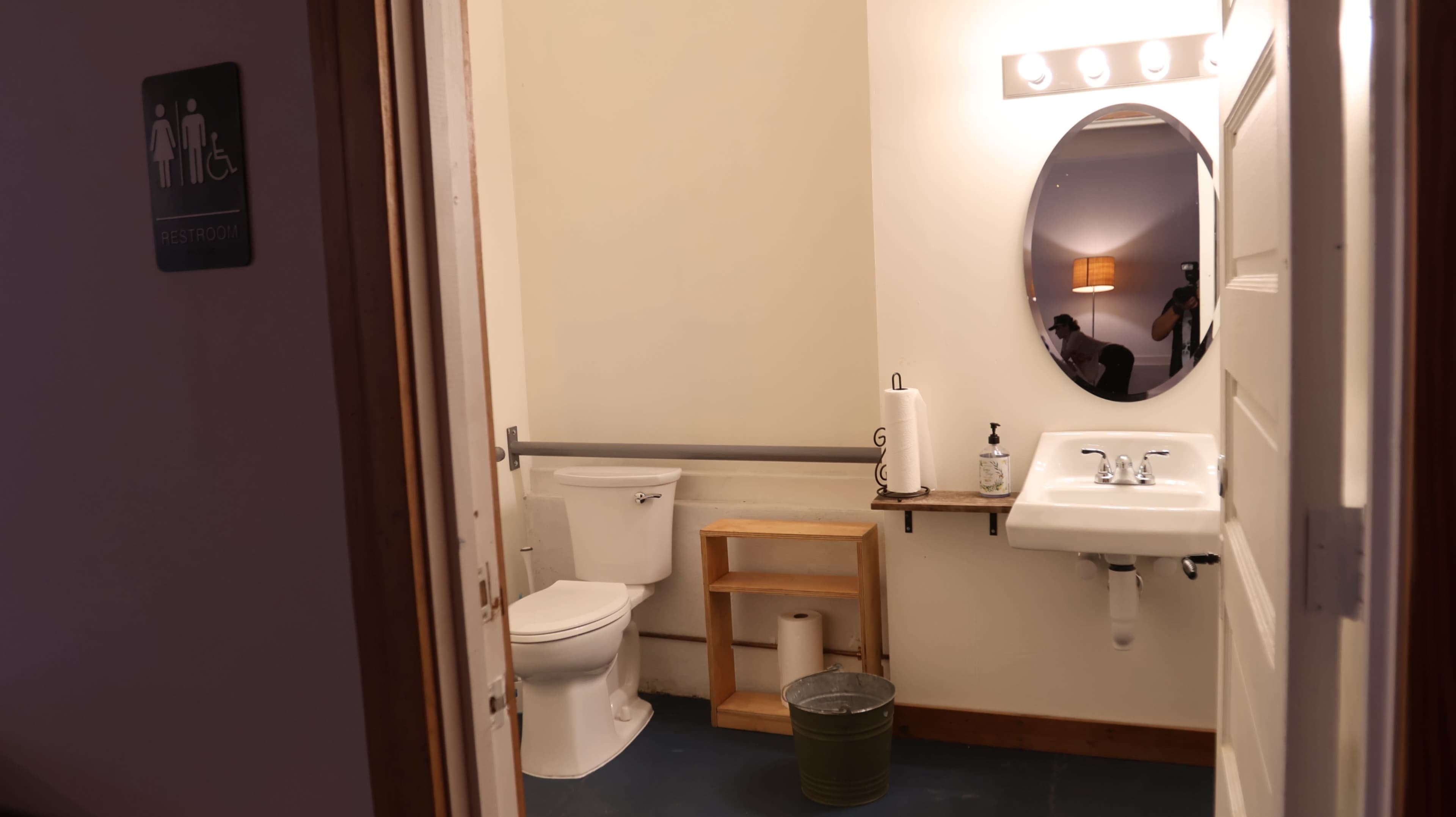The image shows a clean restroom with a toilet, sink, mirror, and a small wooden shelf.