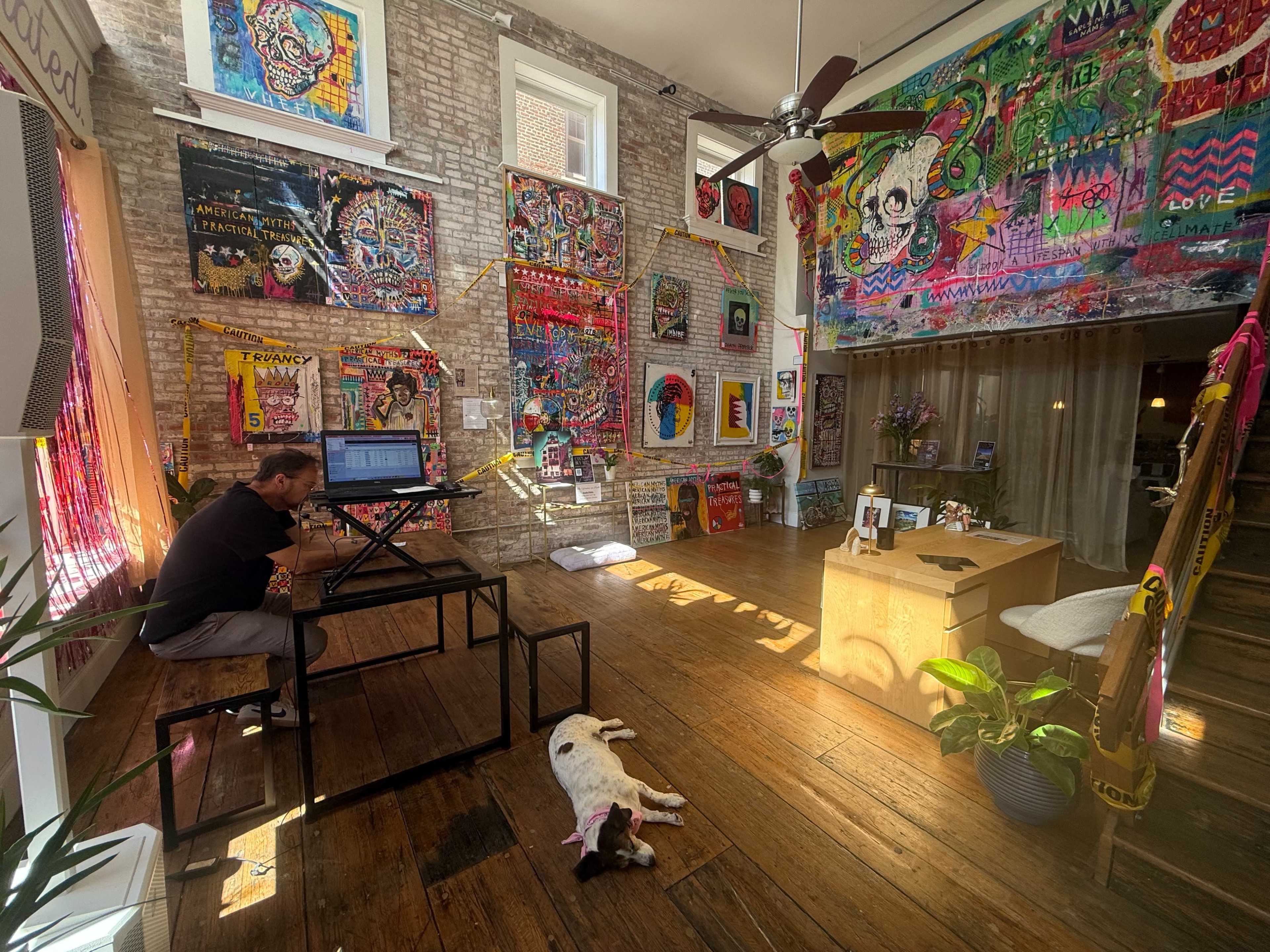 A person works at a desk in an art-filled room with colorful paintings on the walls and a dog lying on the wooden floor.