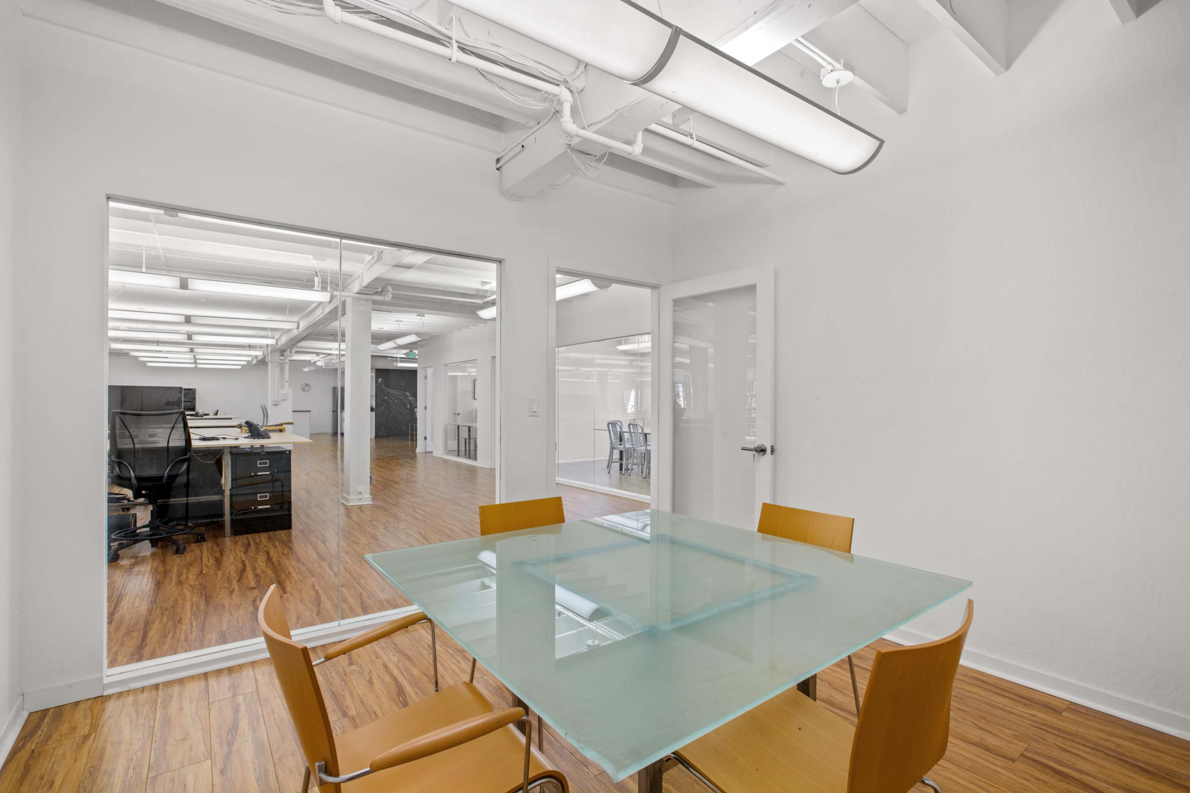 A modern office with a glass conference table and four chairs in a bright room adjacent to a larger workspace.