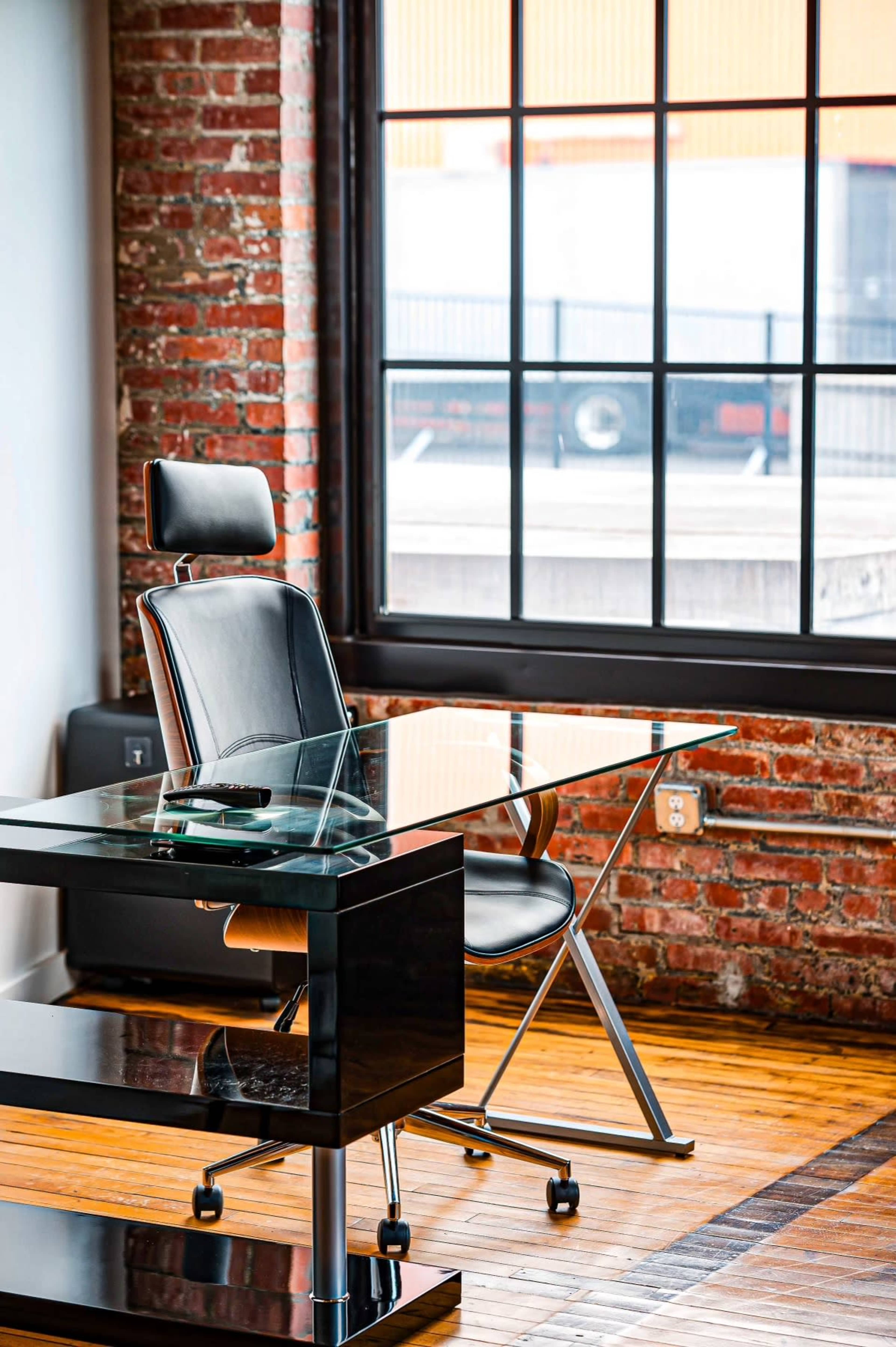 A glass desk and a black leather office chair are positioned near large windows in a room with exposed brick walls.