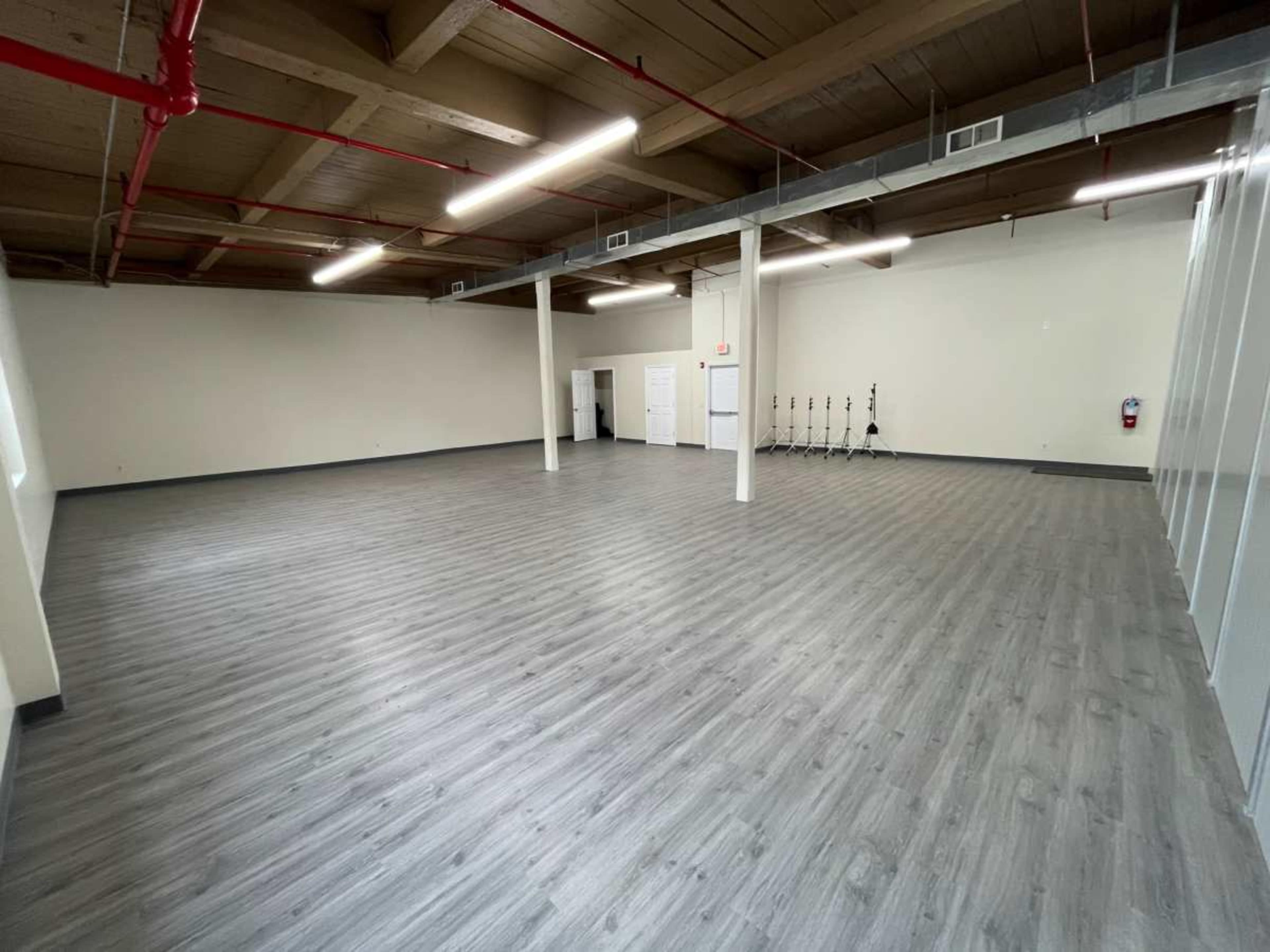 The image shows a spacious, empty room with light-colored wooden flooring and exposed wooden beams on the ceiling.