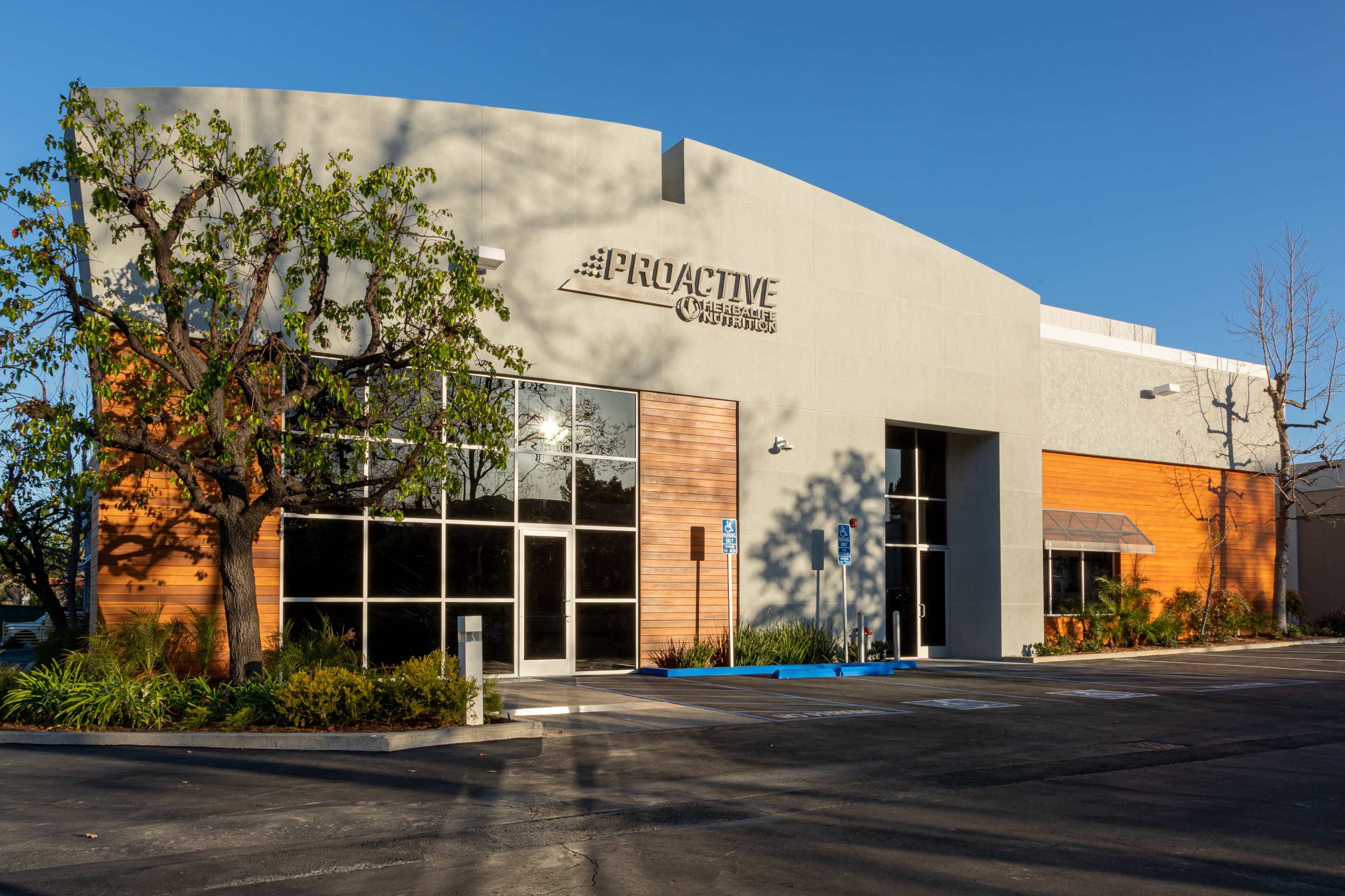 The image shows a modern building with large windows, a curved roof, and a combination of wood and gray exterior, surrounded by landscaped greenery.