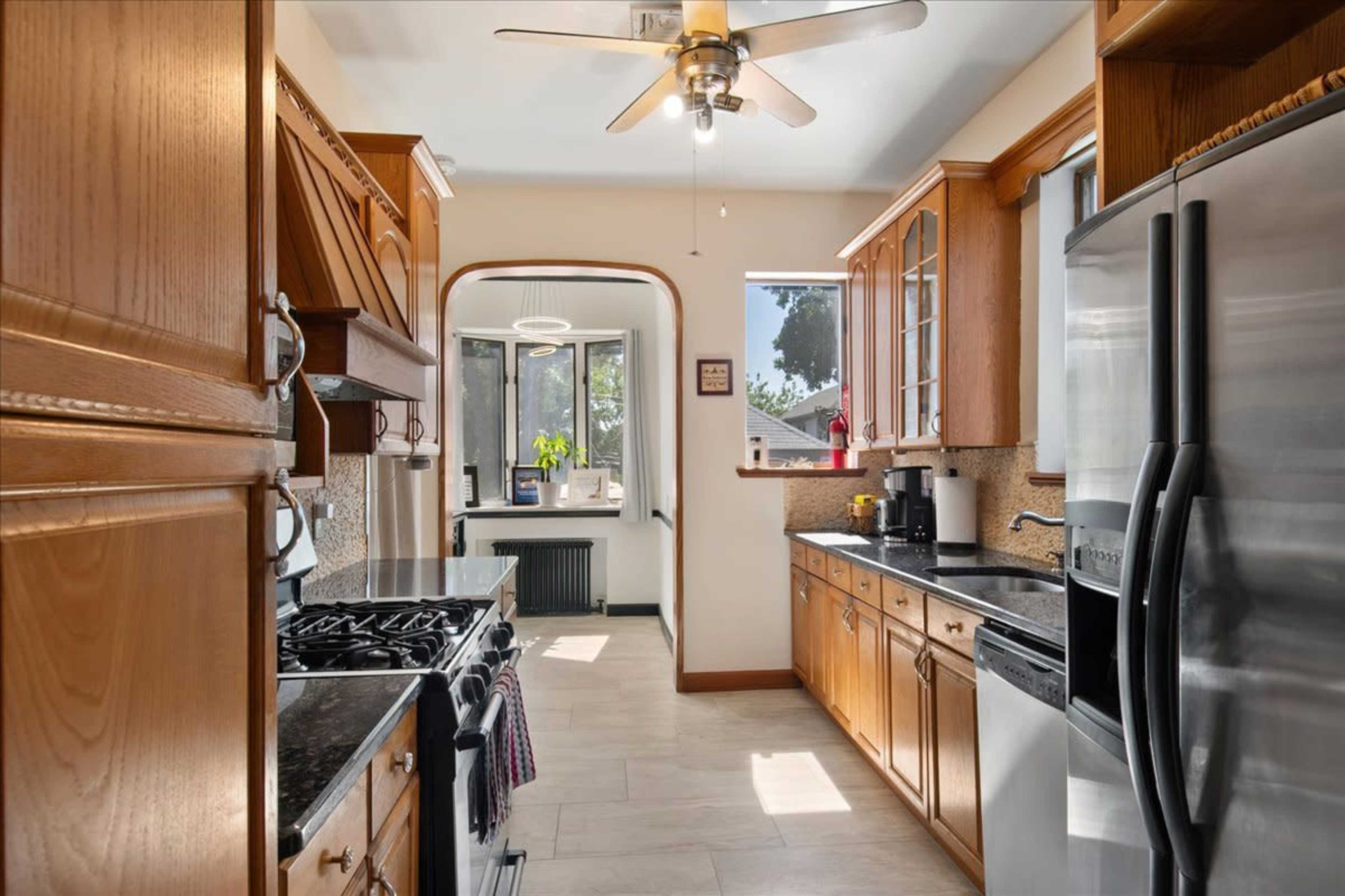 The image shows a kitchen with wooden cabinets, a stainless steel refrigerator, and a stove, illuminated by natural light coming through the windows.