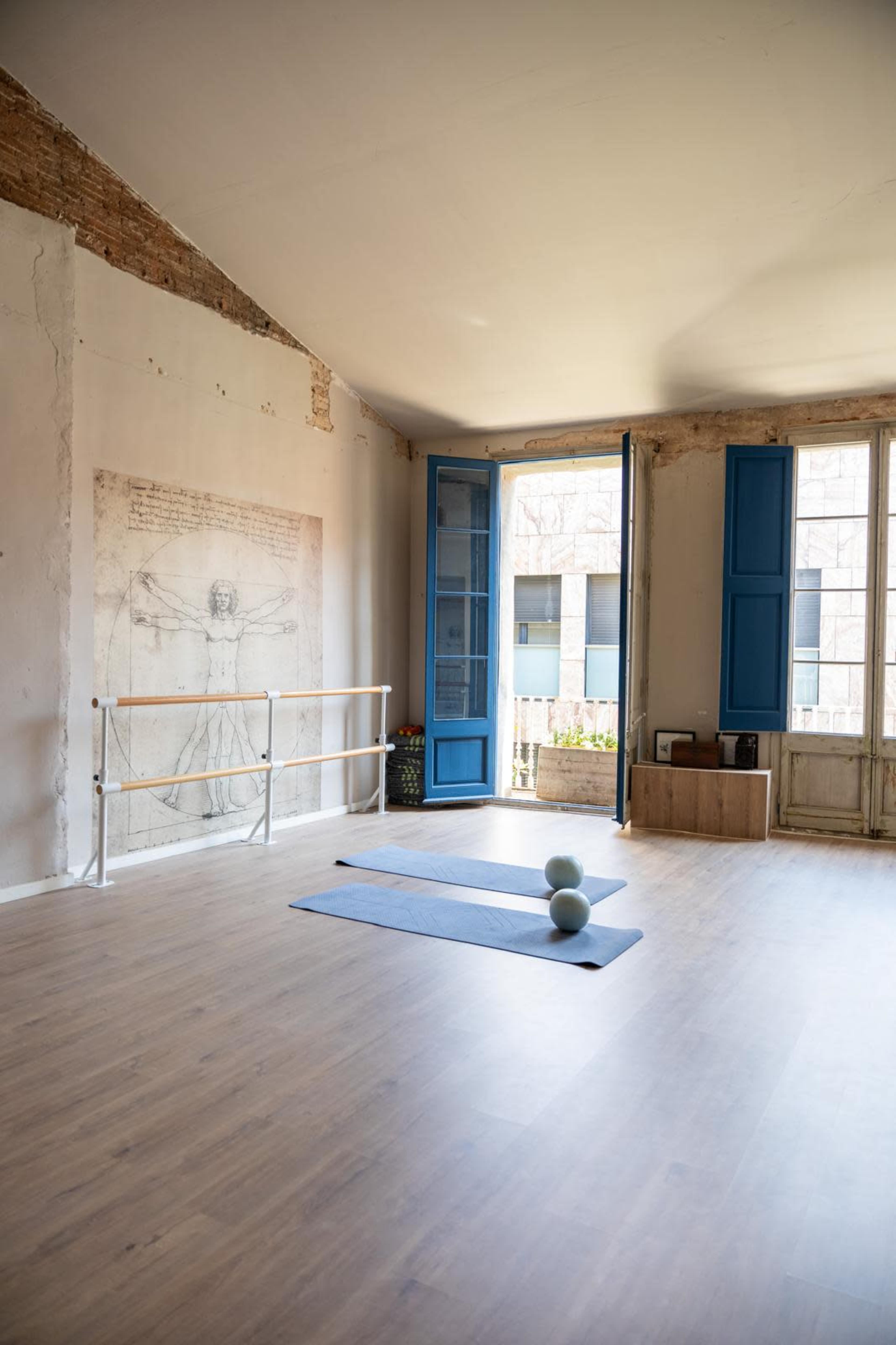 The image shows a bright, spacious room with wooden flooring, two open blue windows, a barre for exercise, and two yoga mats on the floor.
