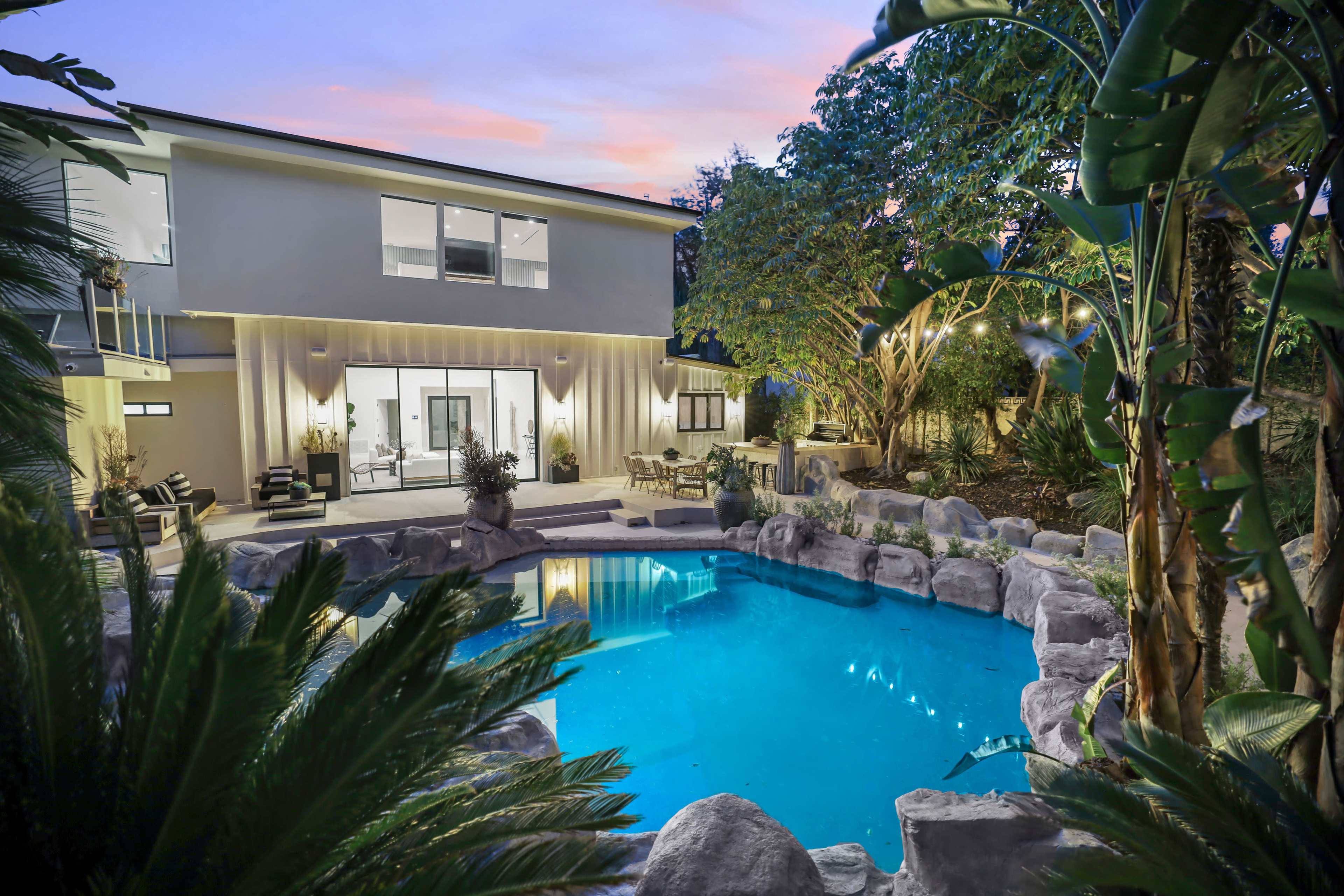 The image shows a modern backyard with a swimming pool surrounded by rocks and tropical plants, featuring a house in the background with large windows.