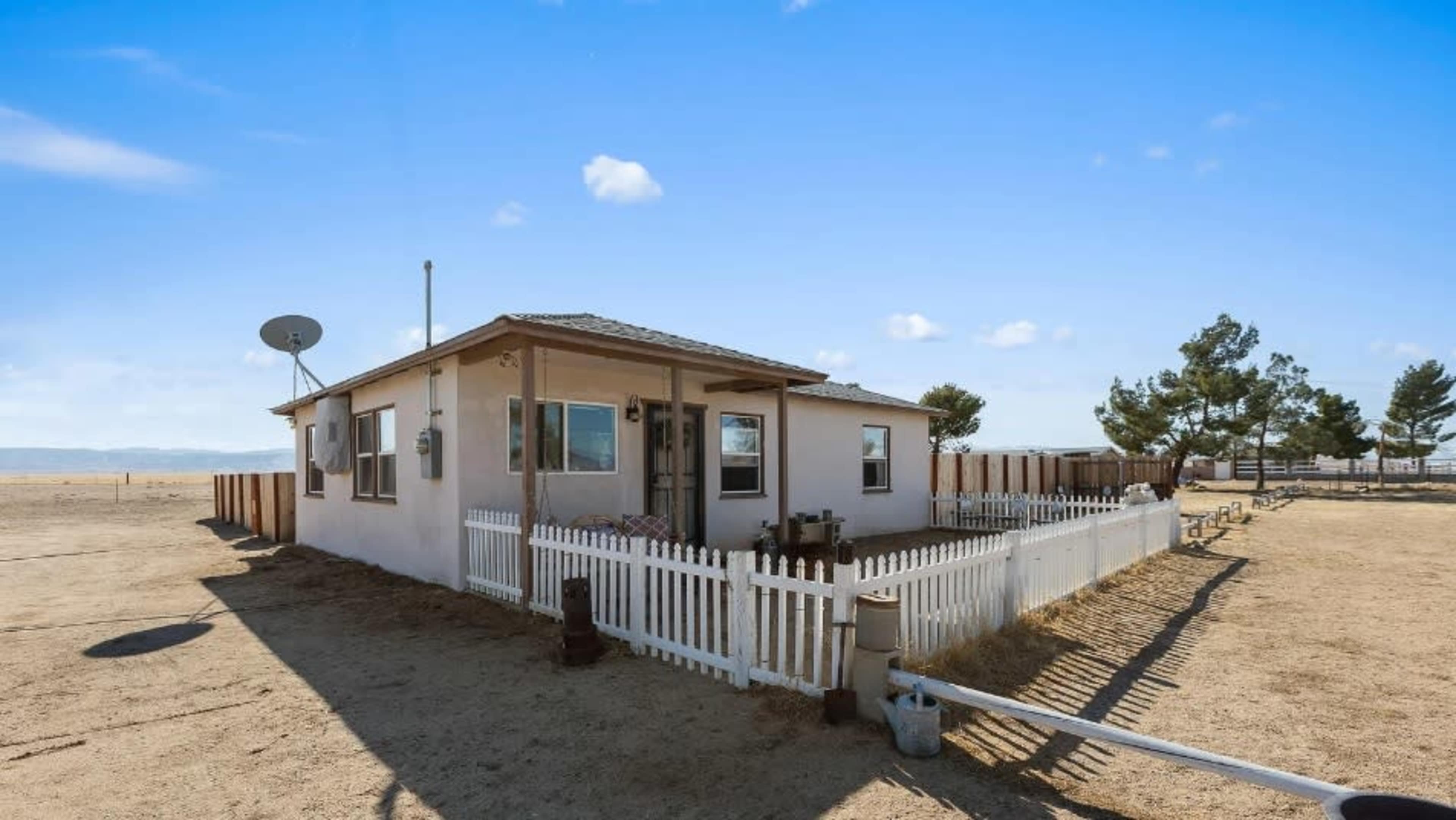 A small house with a white picket fence and a satellite dish is situated in a barren landscape under a blue sky.