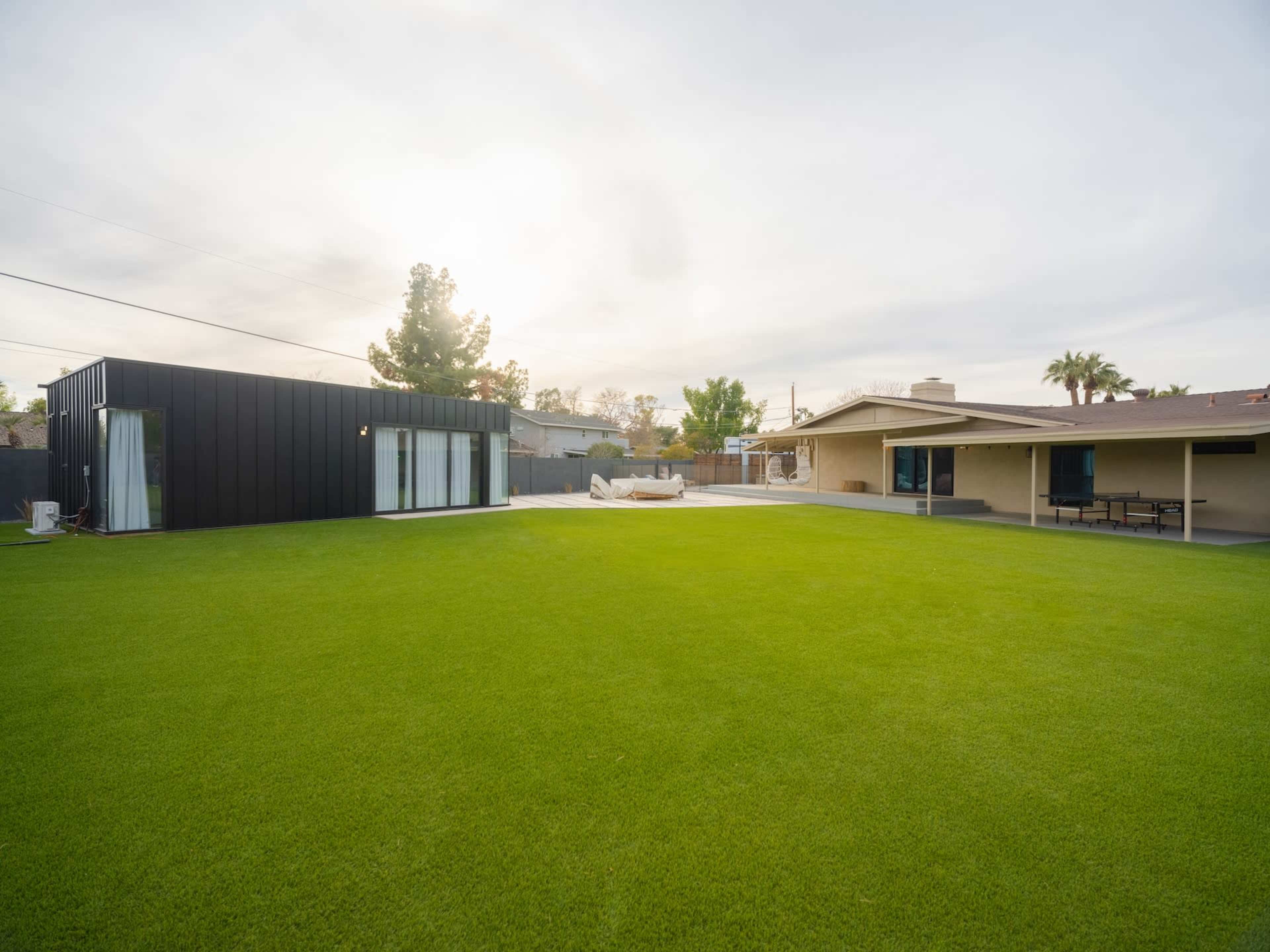 A modern house with a black exterior and large windows sits beside a traditional single-story home, both surrounding a spacious green lawn.