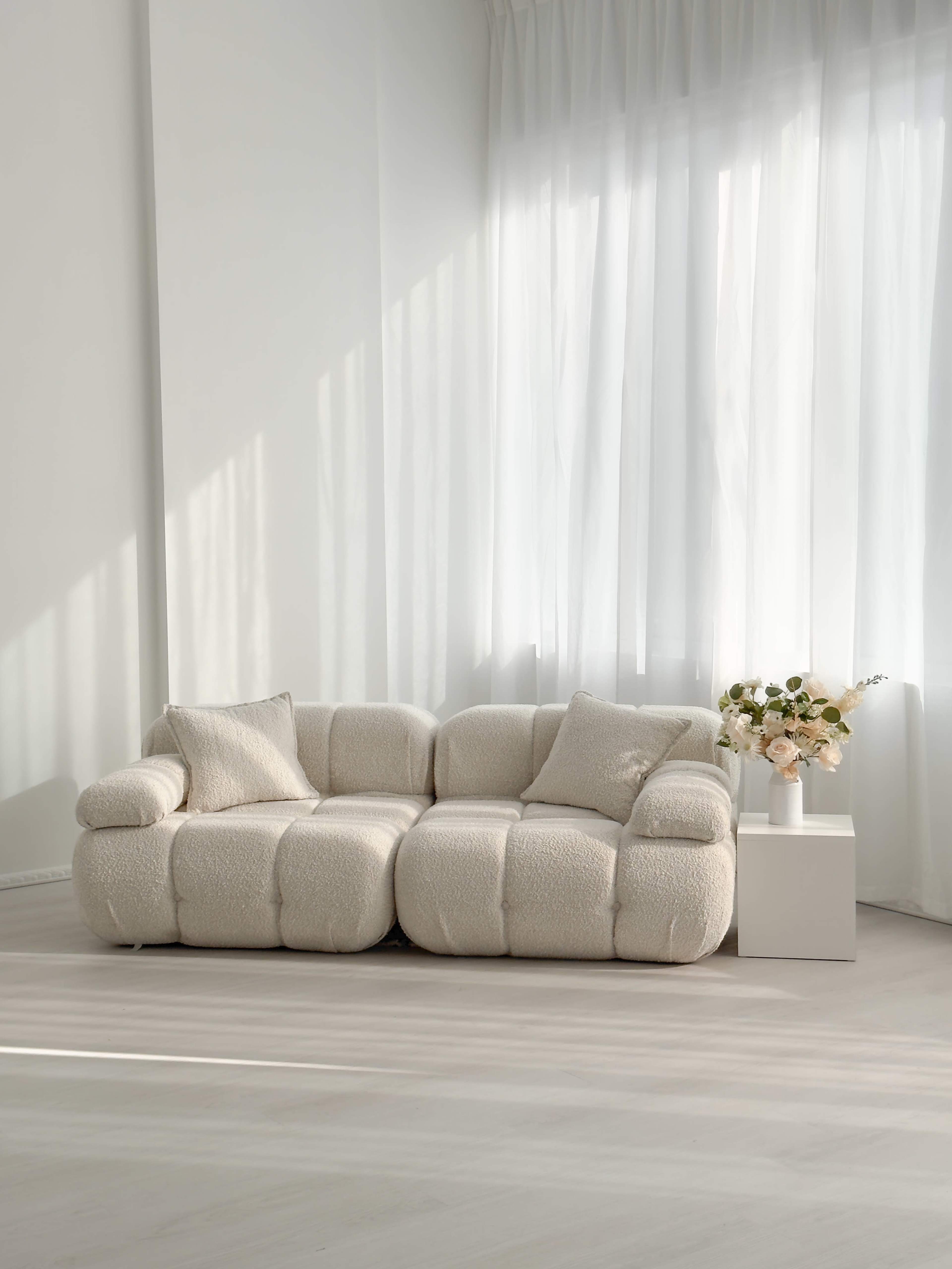 The image shows a light-colored, plush sofa with two cushions next to a white side table holding a vase of flowers, all set against a backdrop of sheer curtains.
