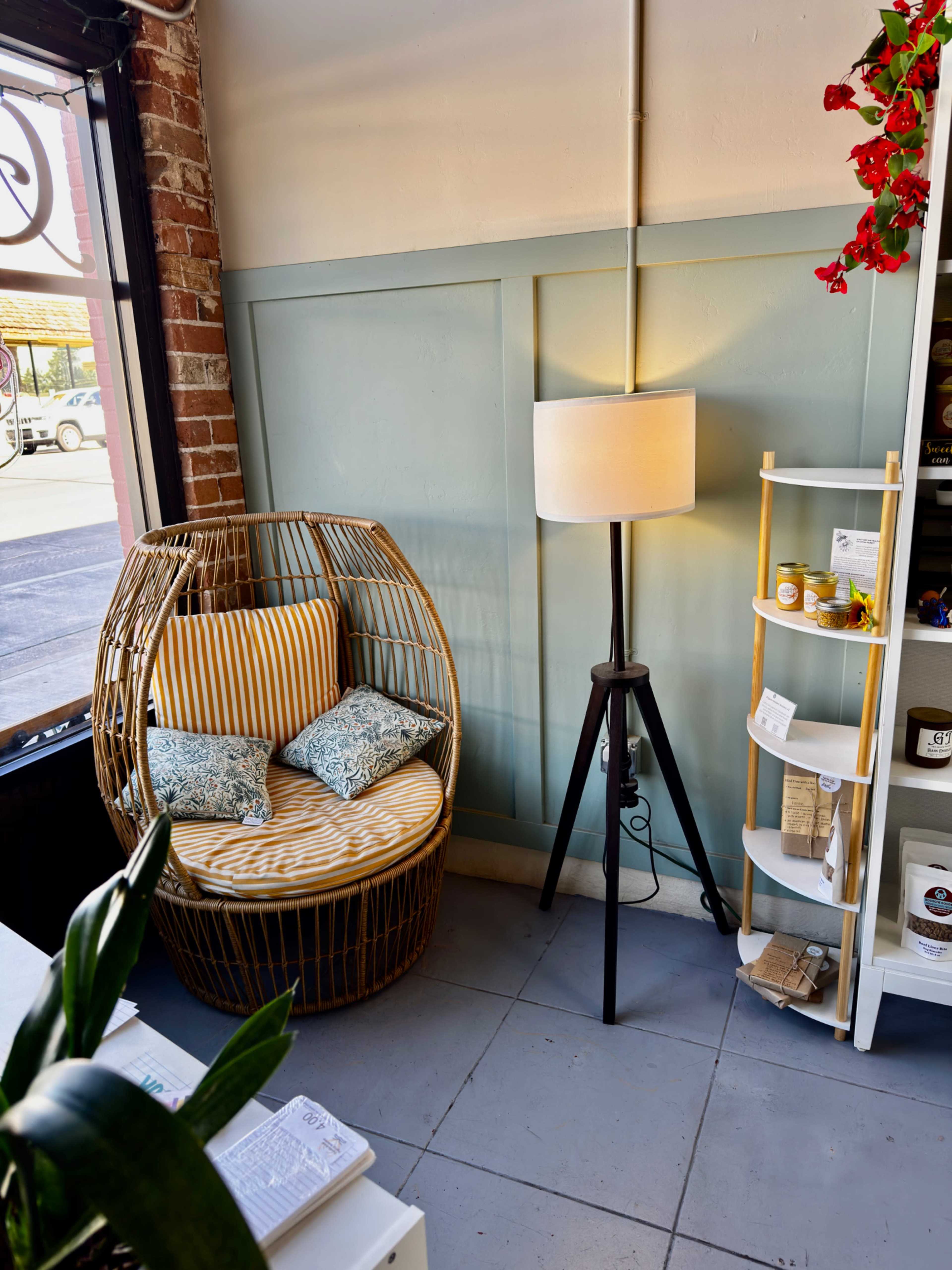 The image shows a cozy corner of a room featuring a round wicker chair with striped cushions, a tall floor lamp, and a white shelving unit displaying various items.