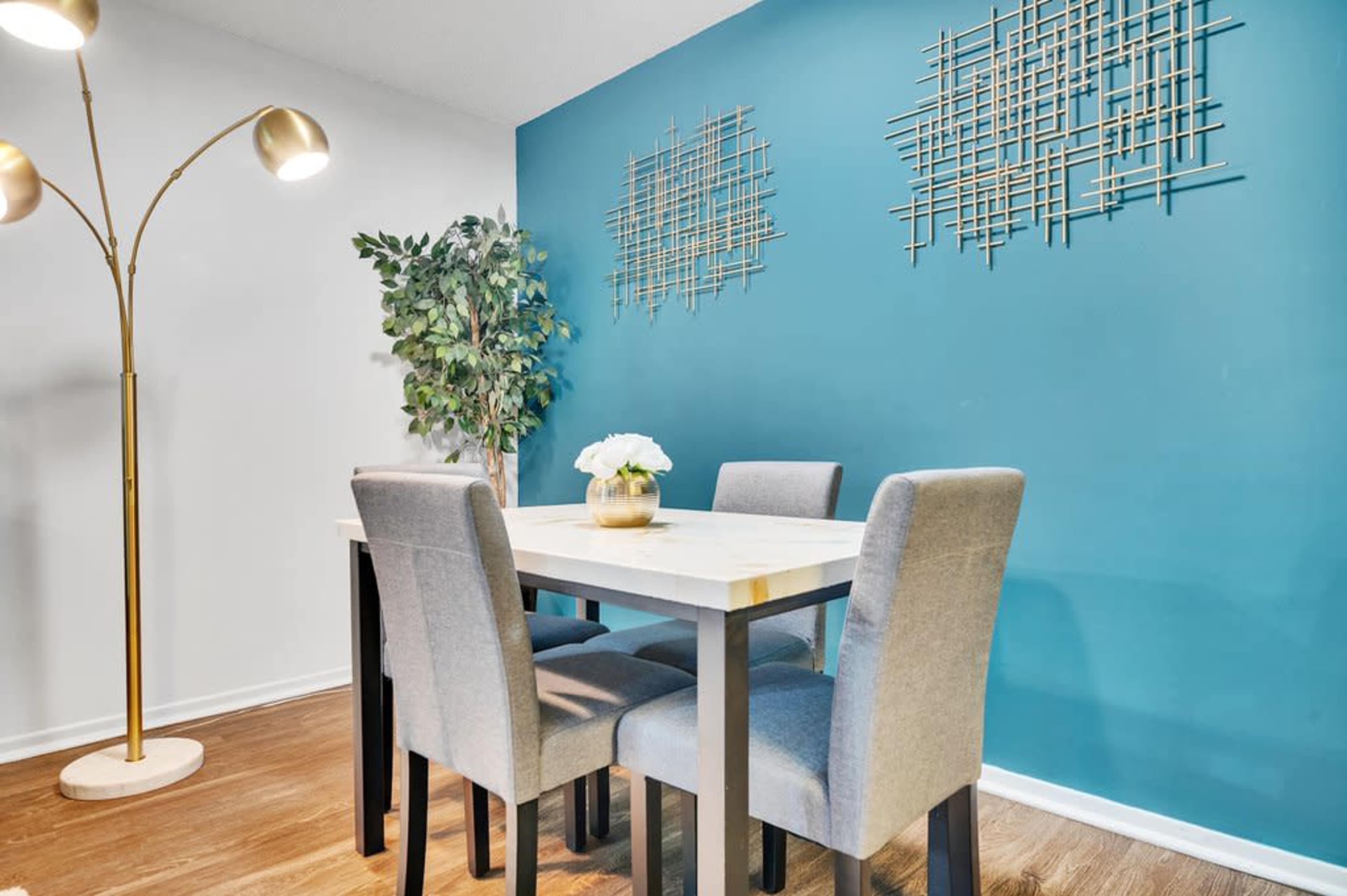 A dining area features a wooden table surrounded by four gray chairs, with a potted plant and decorative wall art on a turquoise wall.