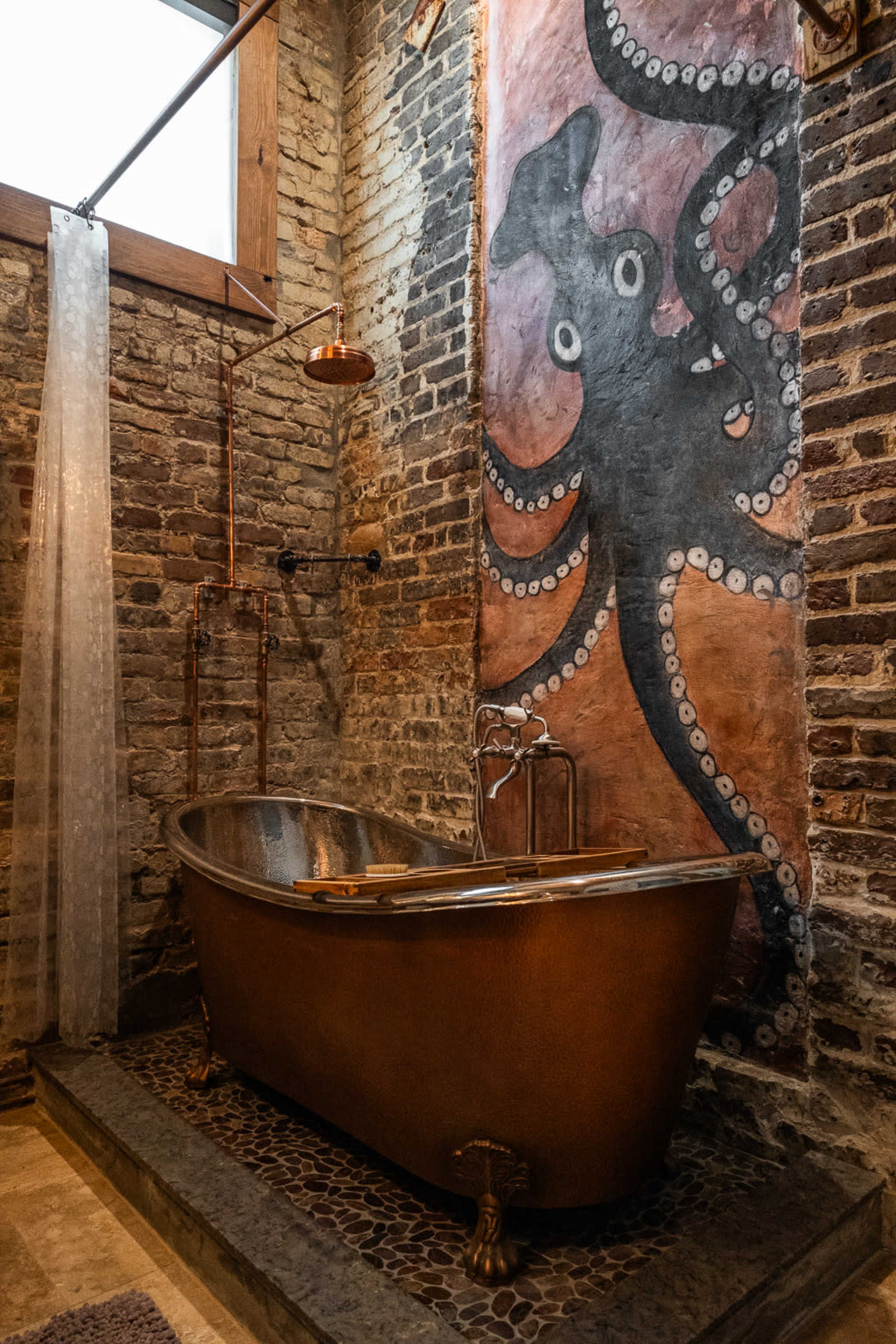 A vintage copper bathtub set against a wall with a mural of an octopus in a rustic bathroom featuring exposed brick and wooden beams.
