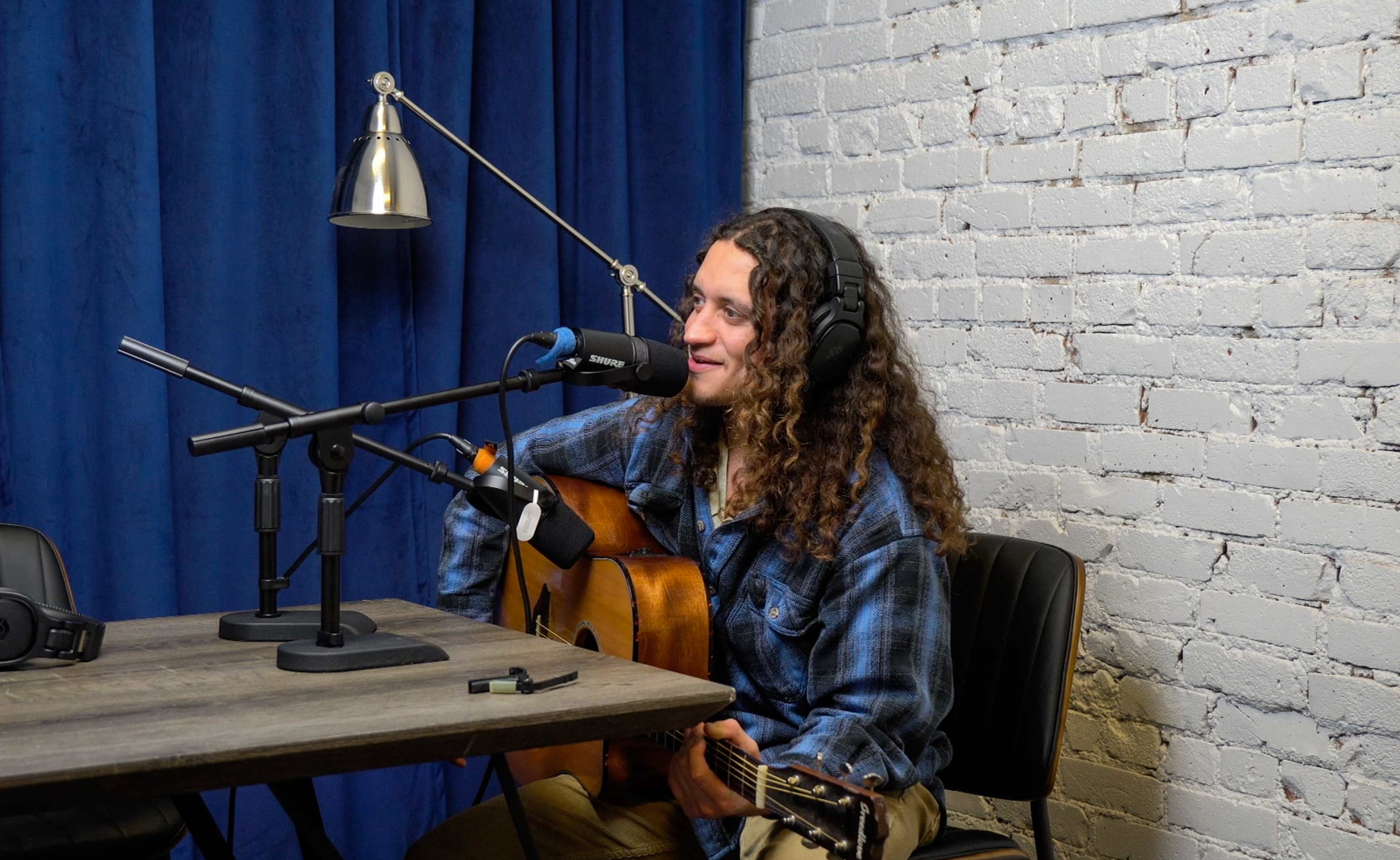 A musician with curly hair plays an acoustic guitar while seated at a table in a recording studio.