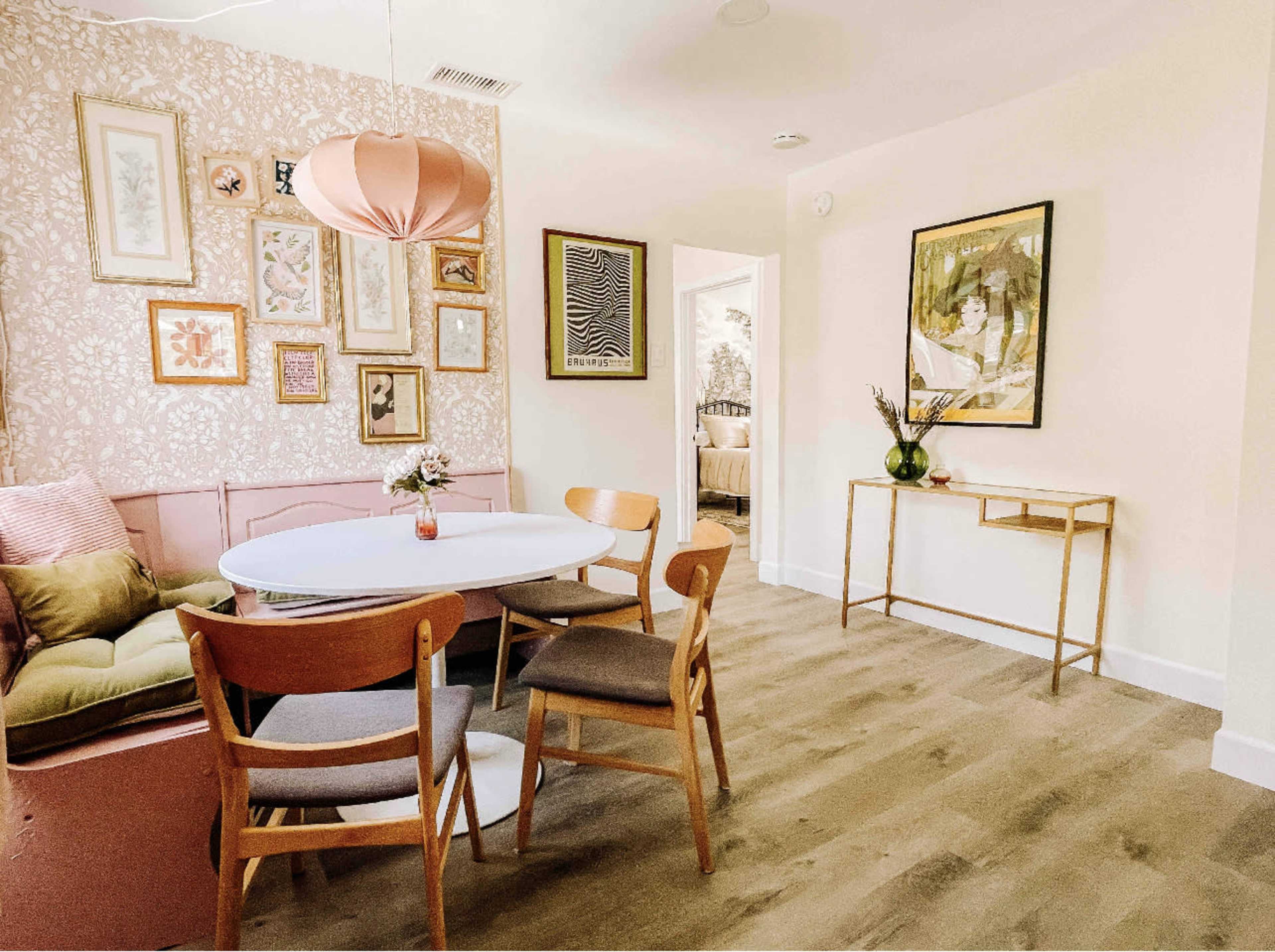 A dining area features a round table surrounded by wooden chairs, with a pink sofa against a floral wallpaper and framed artwork on the walls.