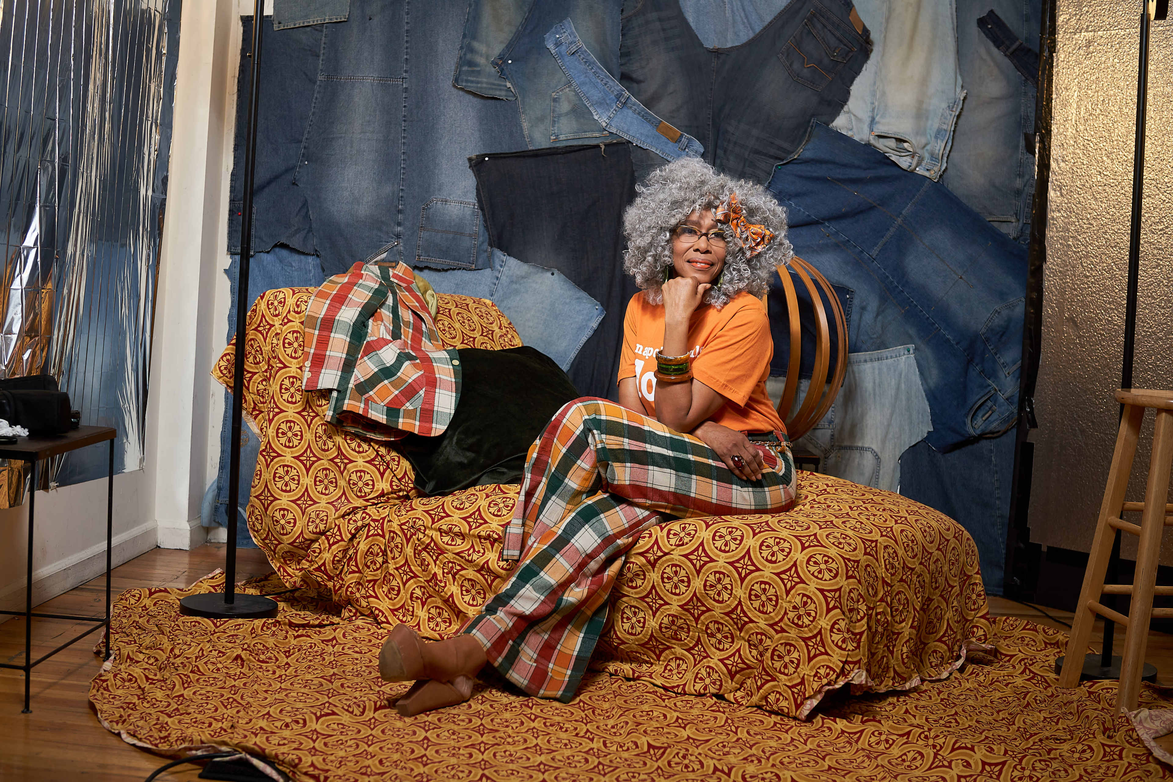 A woman with curly gray hair sits on a patterned bedspread in front of a backdrop made of denim fabric.