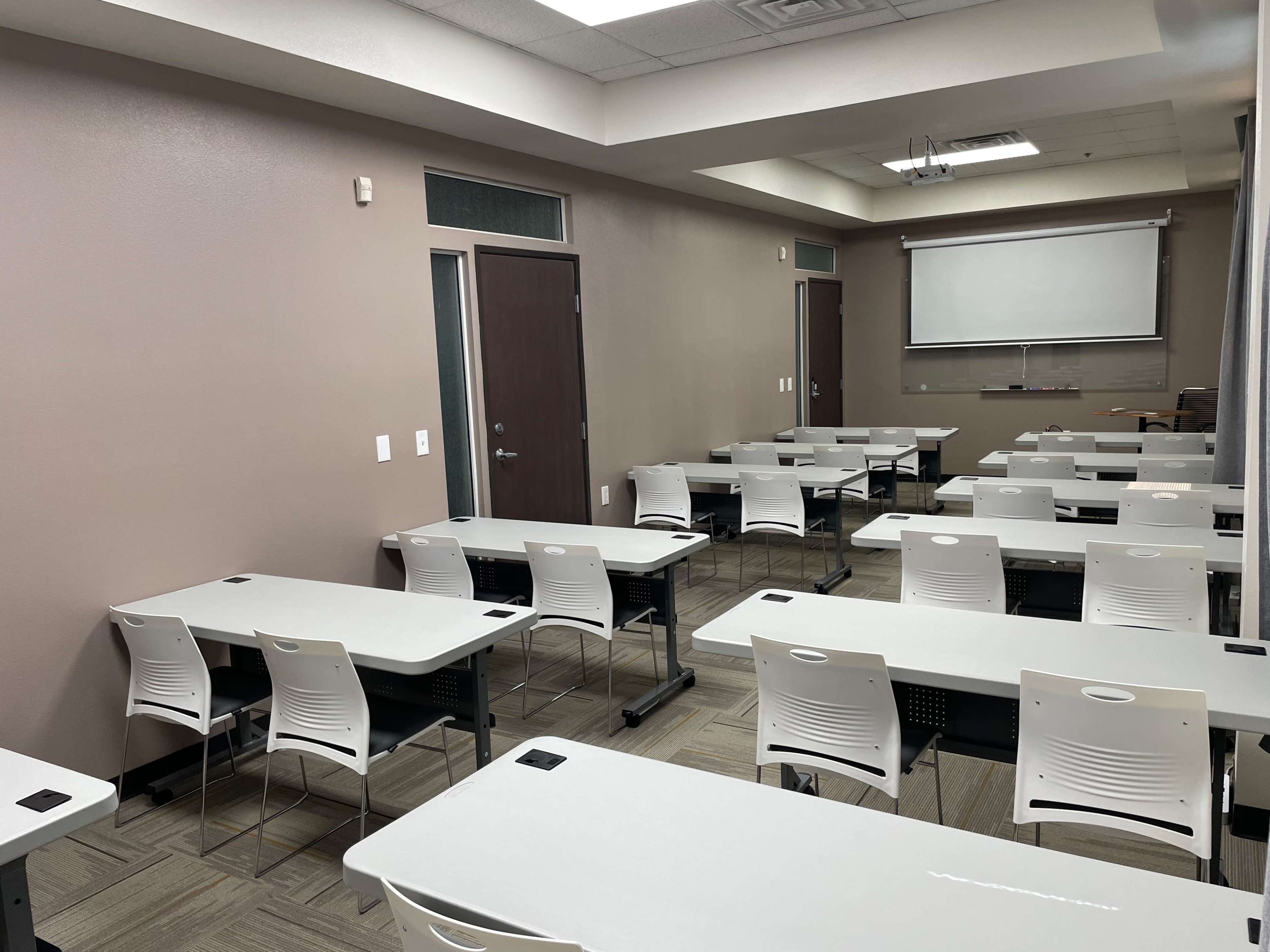 The image shows a classroom with several tables and chairs arranged in rows, and a whiteboard at the front.