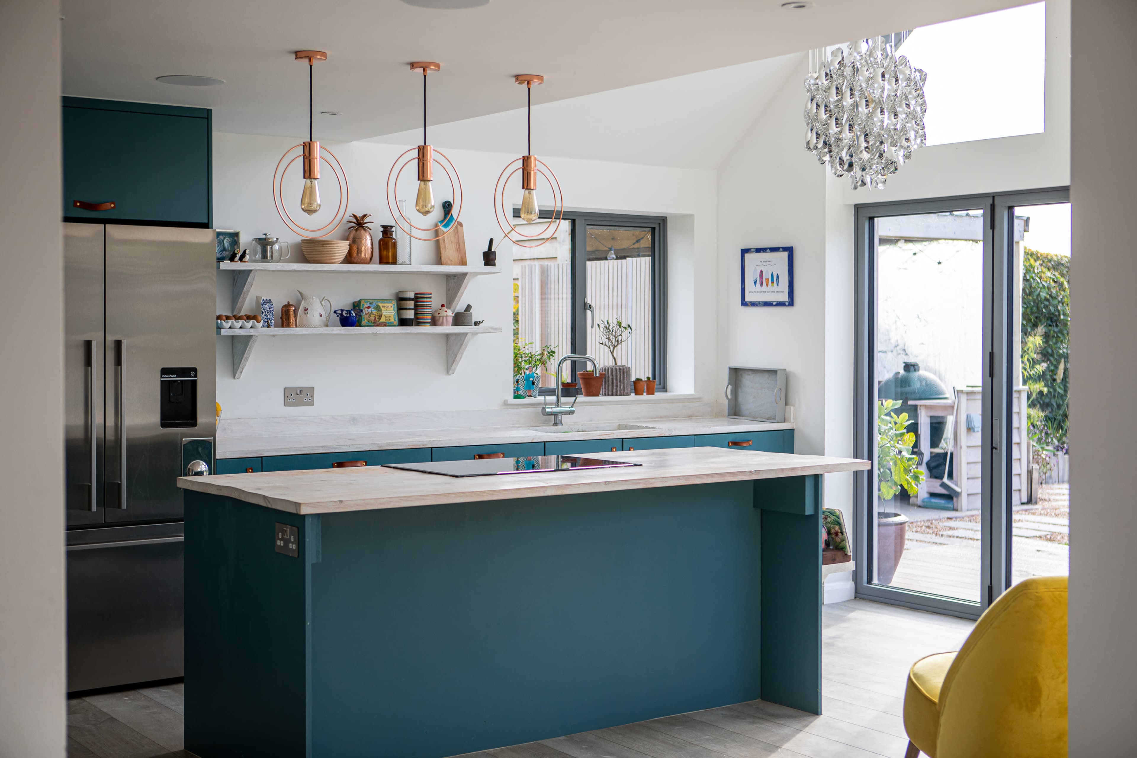 A modern kitchen with teal cabinetry, a central island, and pendant lights hanging above it, featuring a mixture of open shelves and stainless steel appliances.