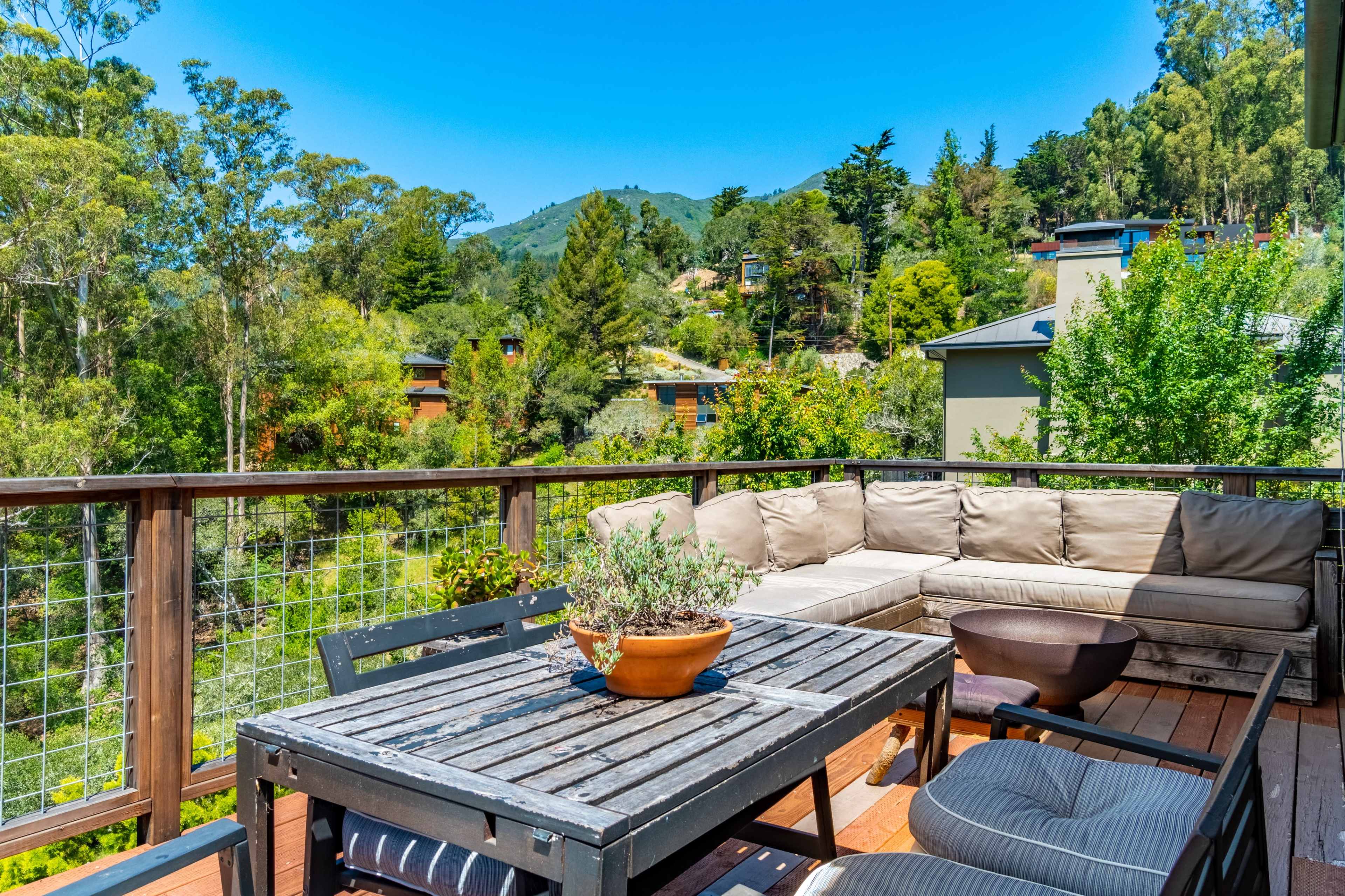 A wooden deck with a seating area and a table overlooks a green landscape with trees and houses.