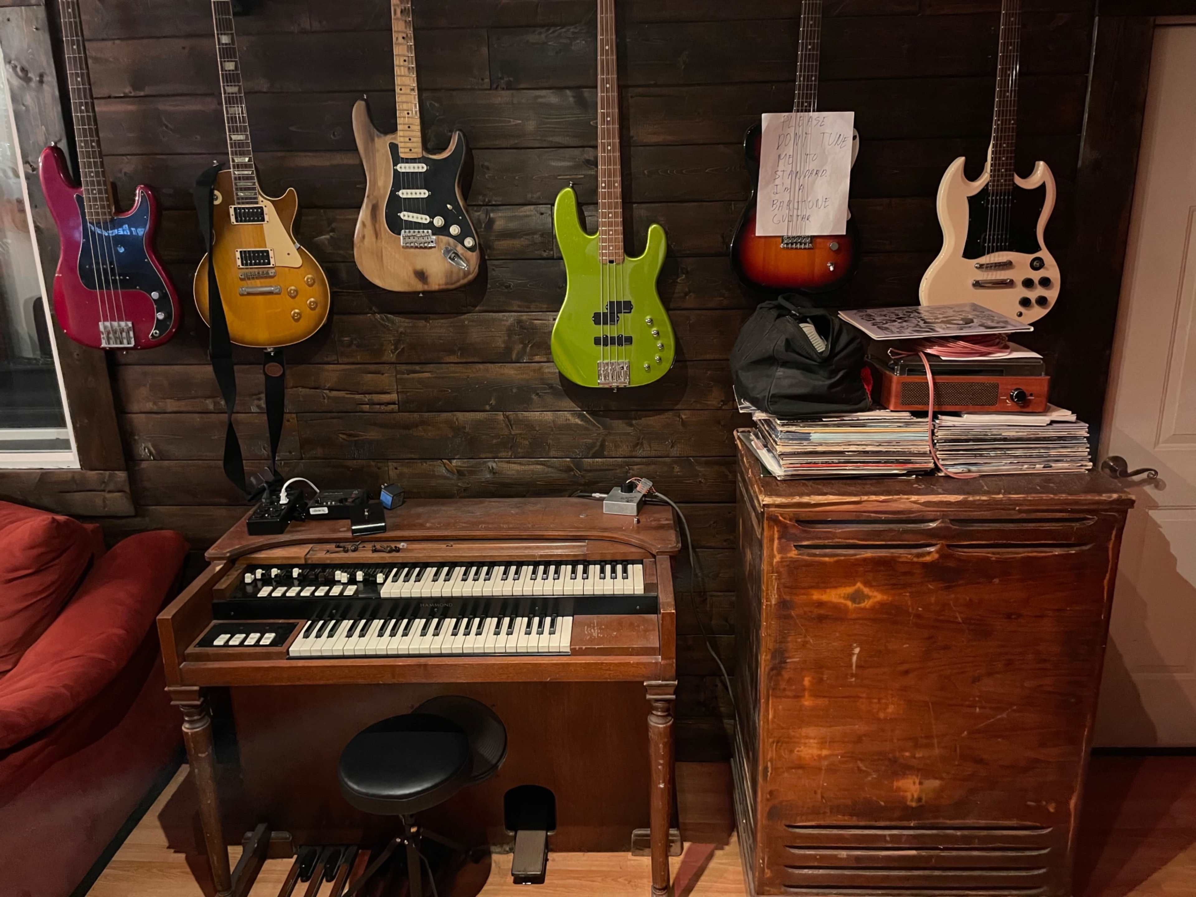 The image shows a vintage organ and a stack of records next to a wooden cabinet, with several electric guitars hanging on the wall behind them.