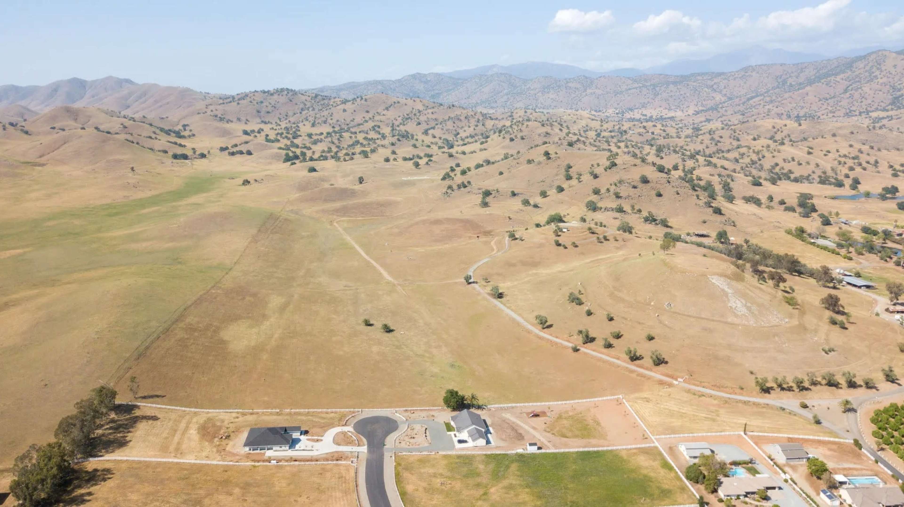 The image features a vast, dry landscape with rolling hills, sparse vegetation, and a few scattered homes along a winding road.