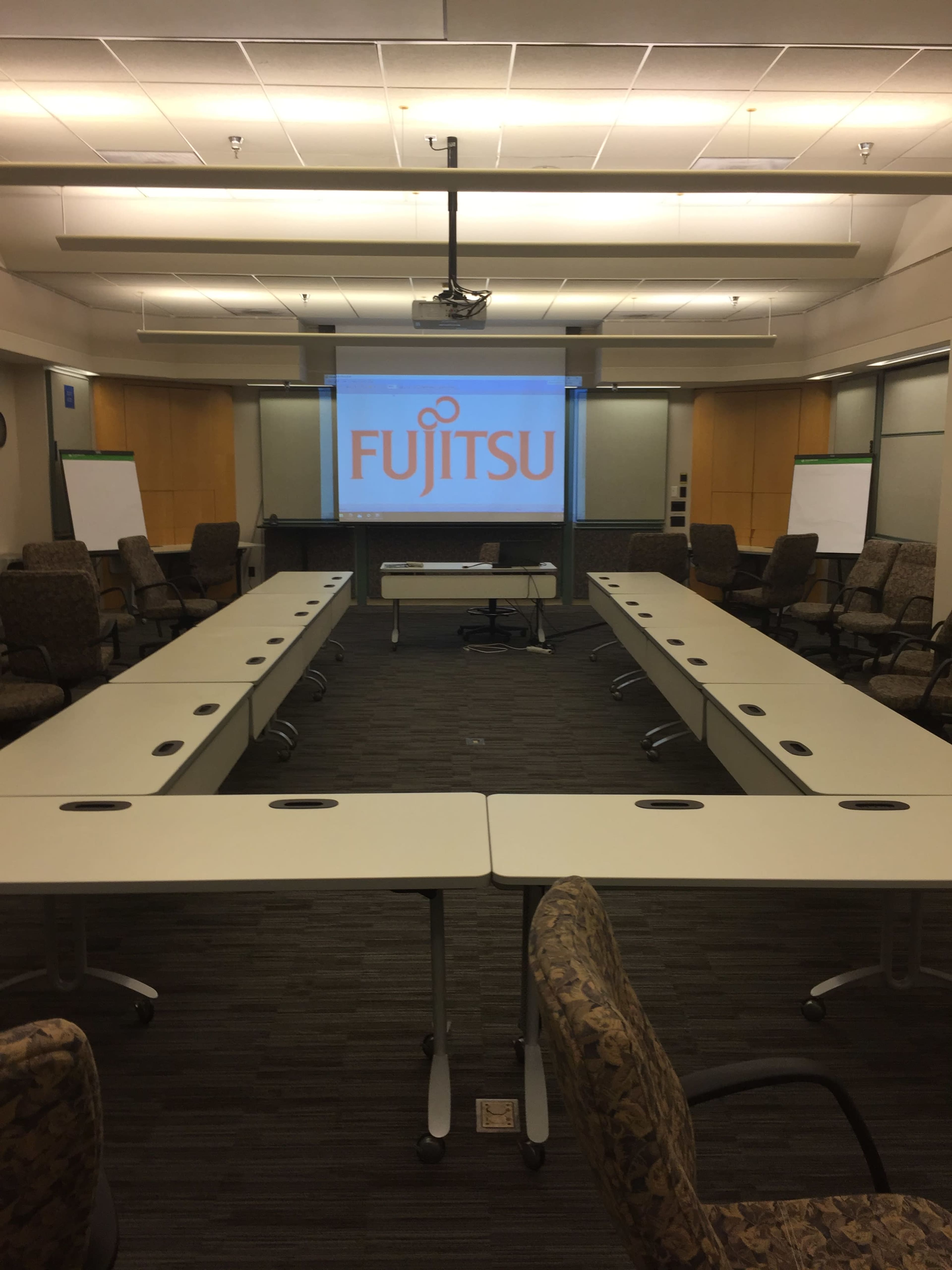 A conference room is organized with tables arranged in a U-shape, featuring a projector screen displaying the Fujitsu logo at the front.